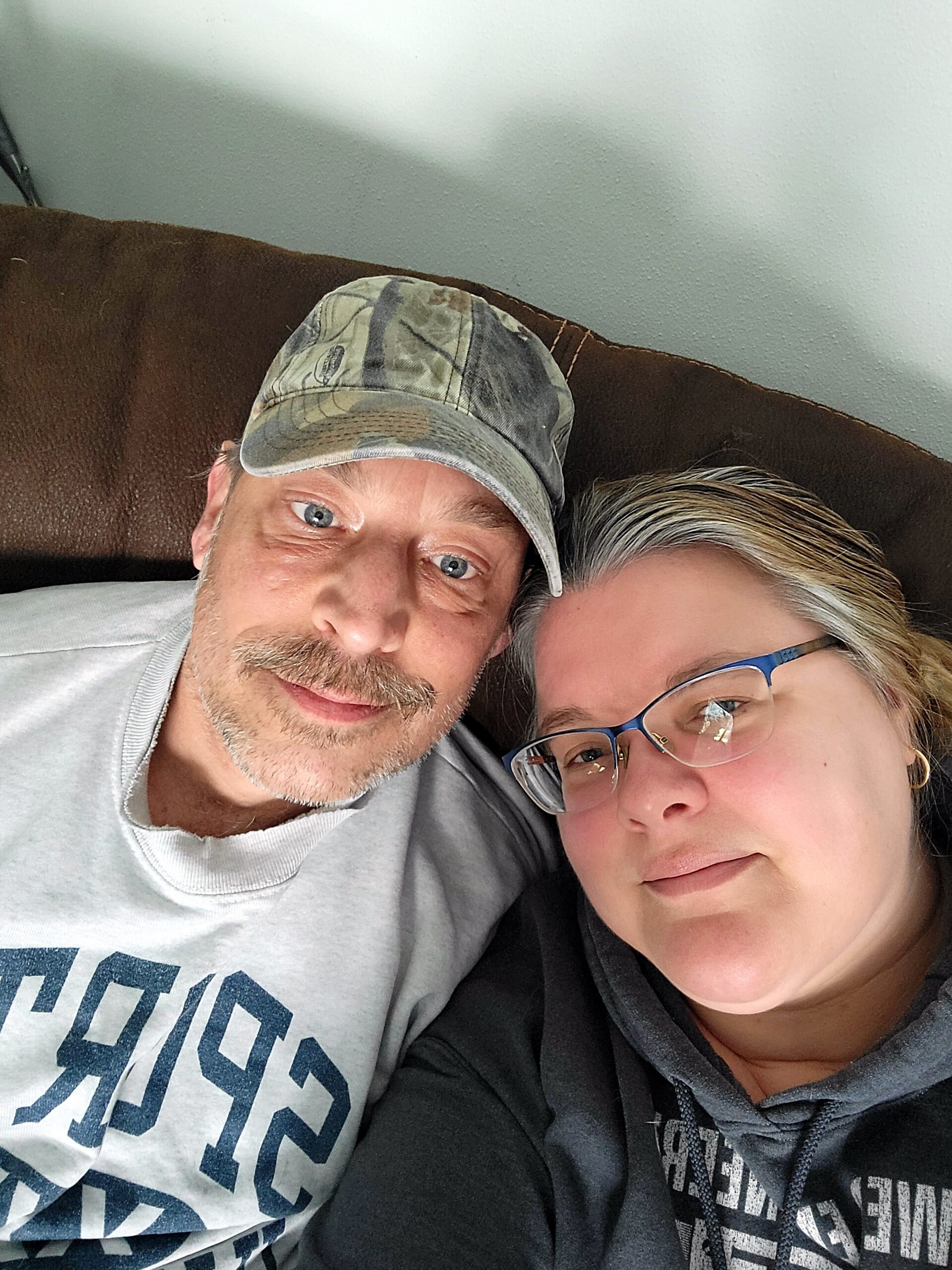 Close-up selfie of a man in a camo cap and a gray shirt with a woman wearing glasses resting on a brown couch beside him.