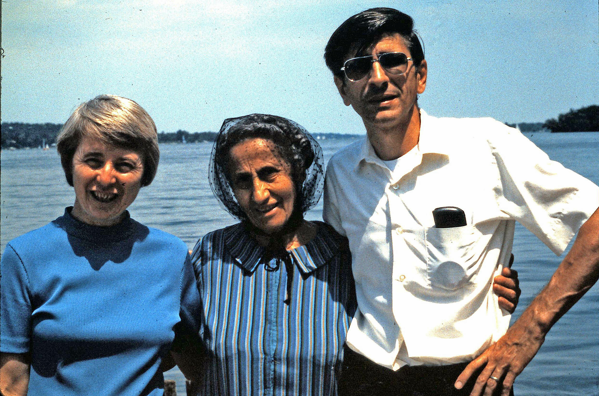 Three adults posing together by a lake: woman in blue sweater on the left, woman in a striped dress with a veil in the middle, man in a white shirt with sunglasses on the right, all smiling at the camera.
