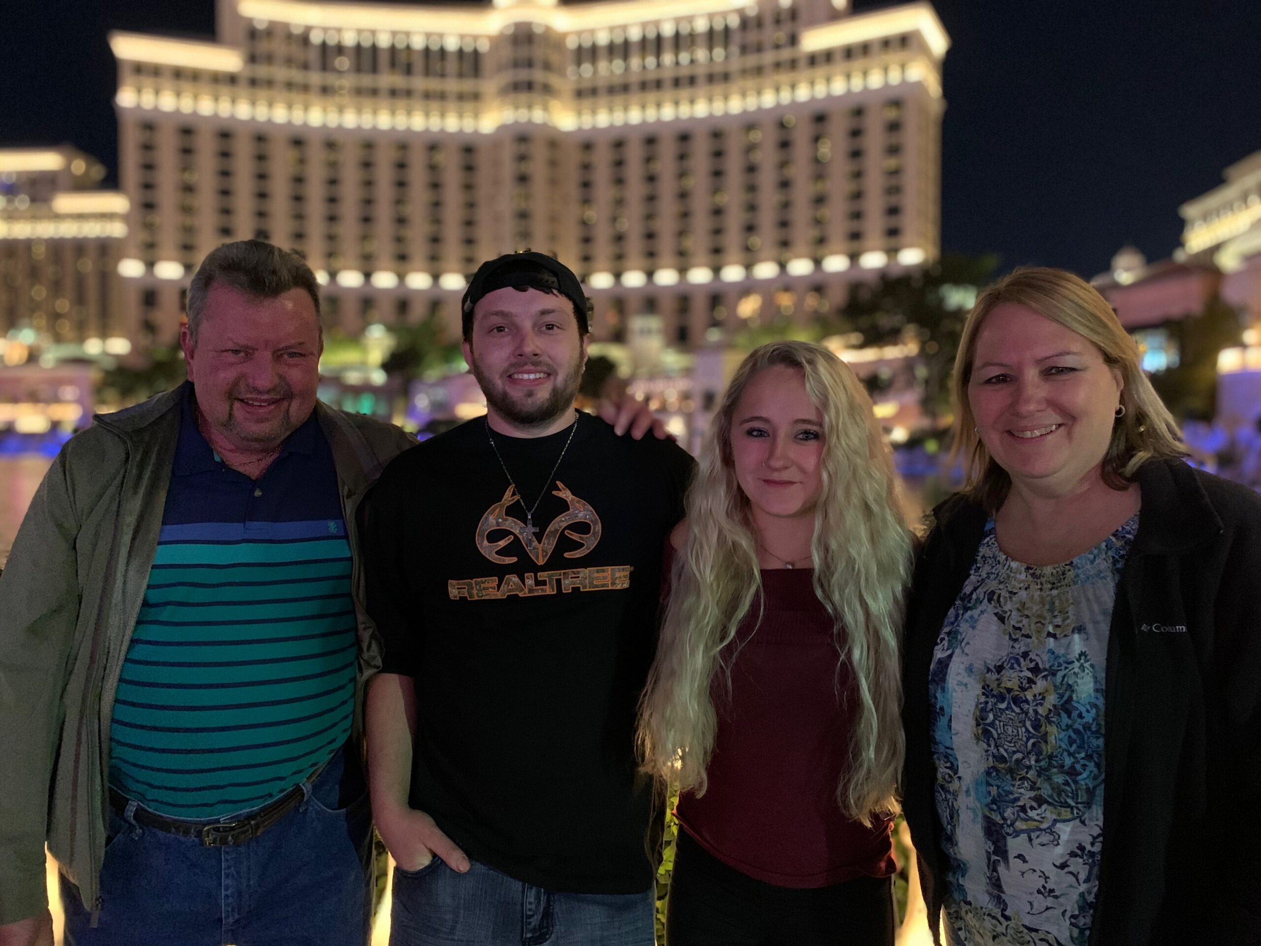 Four adults stand close together at night, smiling, with a brightly lit hotel/casino building in the background.