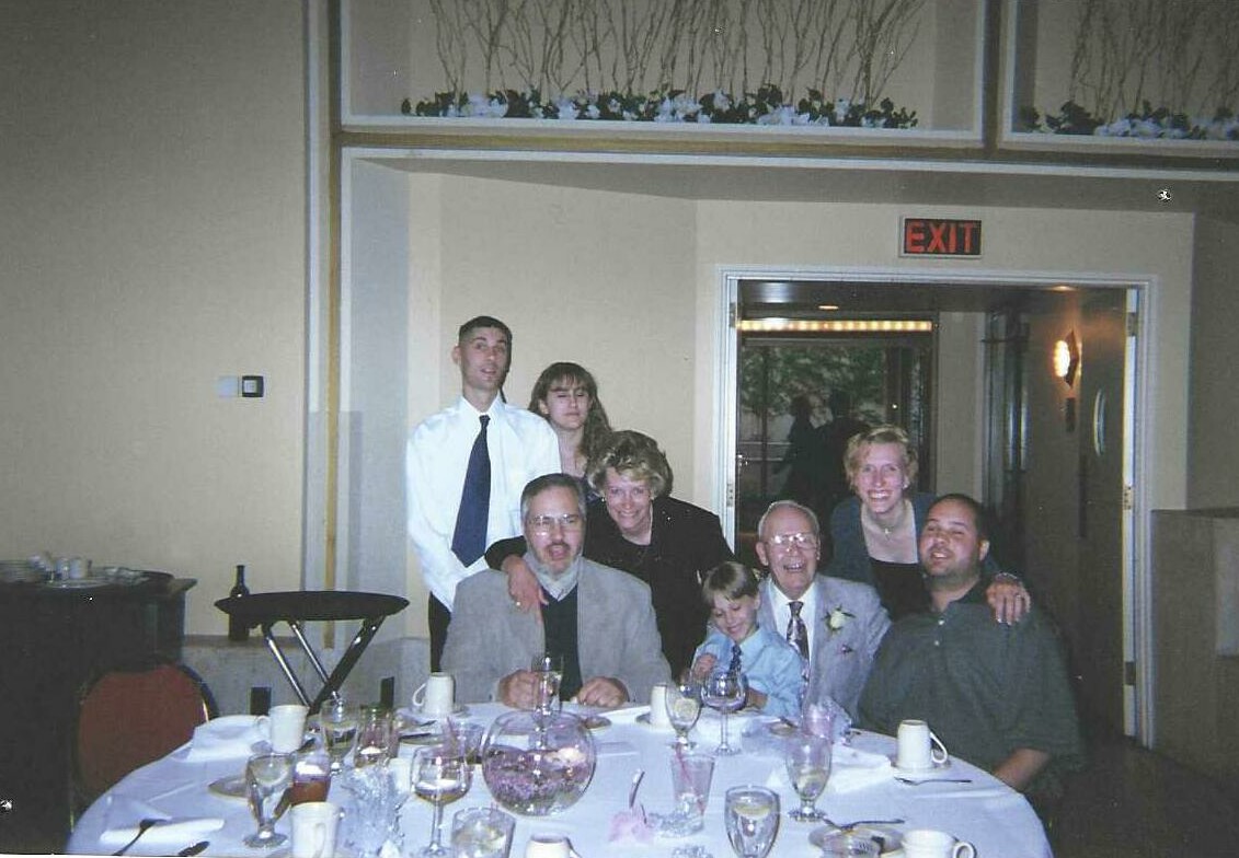 Group of eight people gathered around a round banquet table, posing for a photo at a formal event near an exit sign in the background.