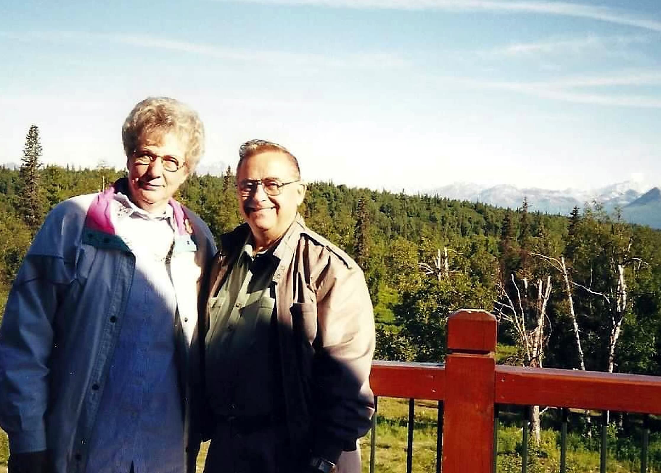 Two smiling men posing together outdoors on a wooden railing with trees and mountains in the background.