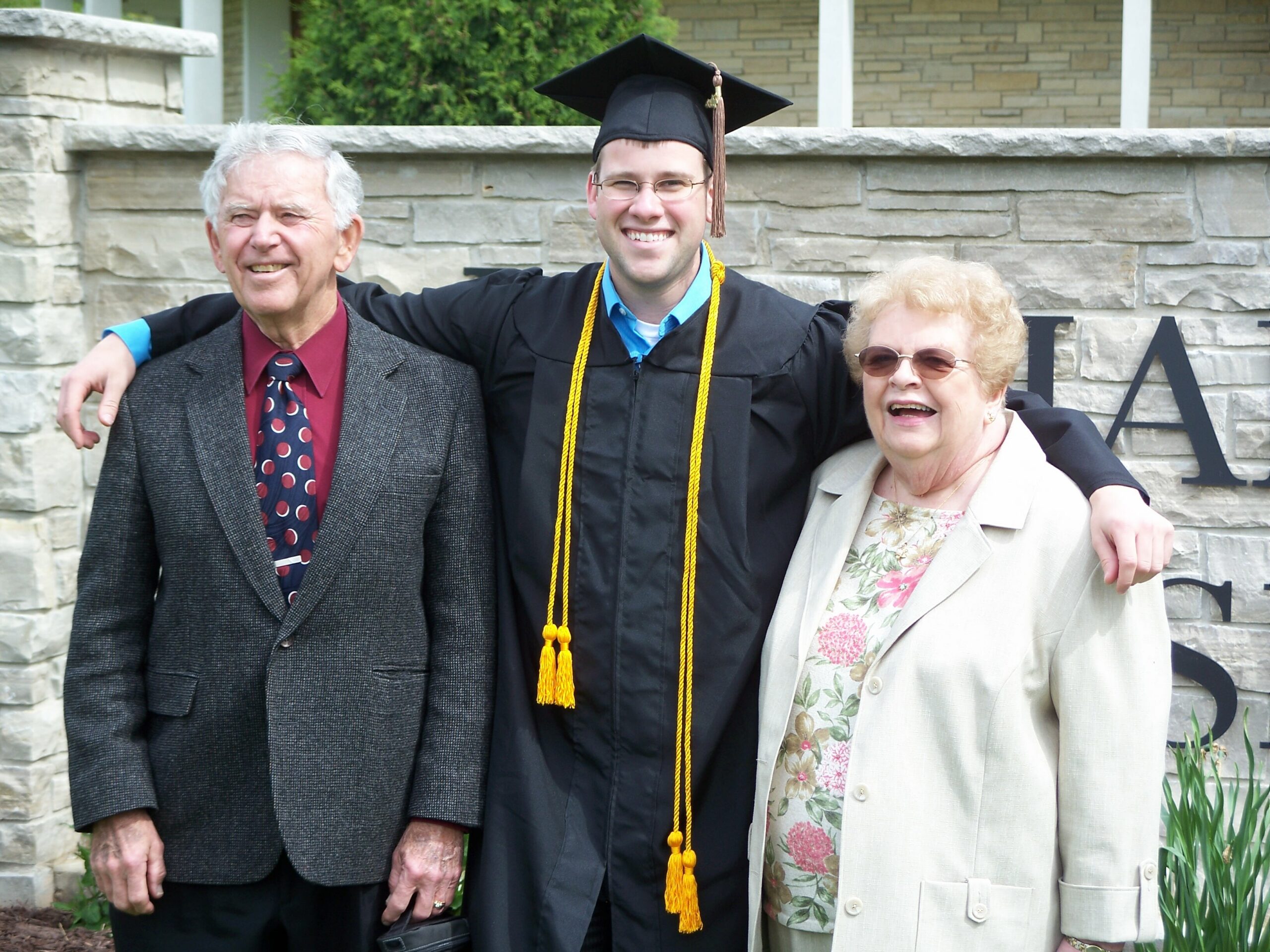 Graduate in cap and gown with yellow cords, flanked by an elderly man and woman, posing outdoors in front of a stone wall.