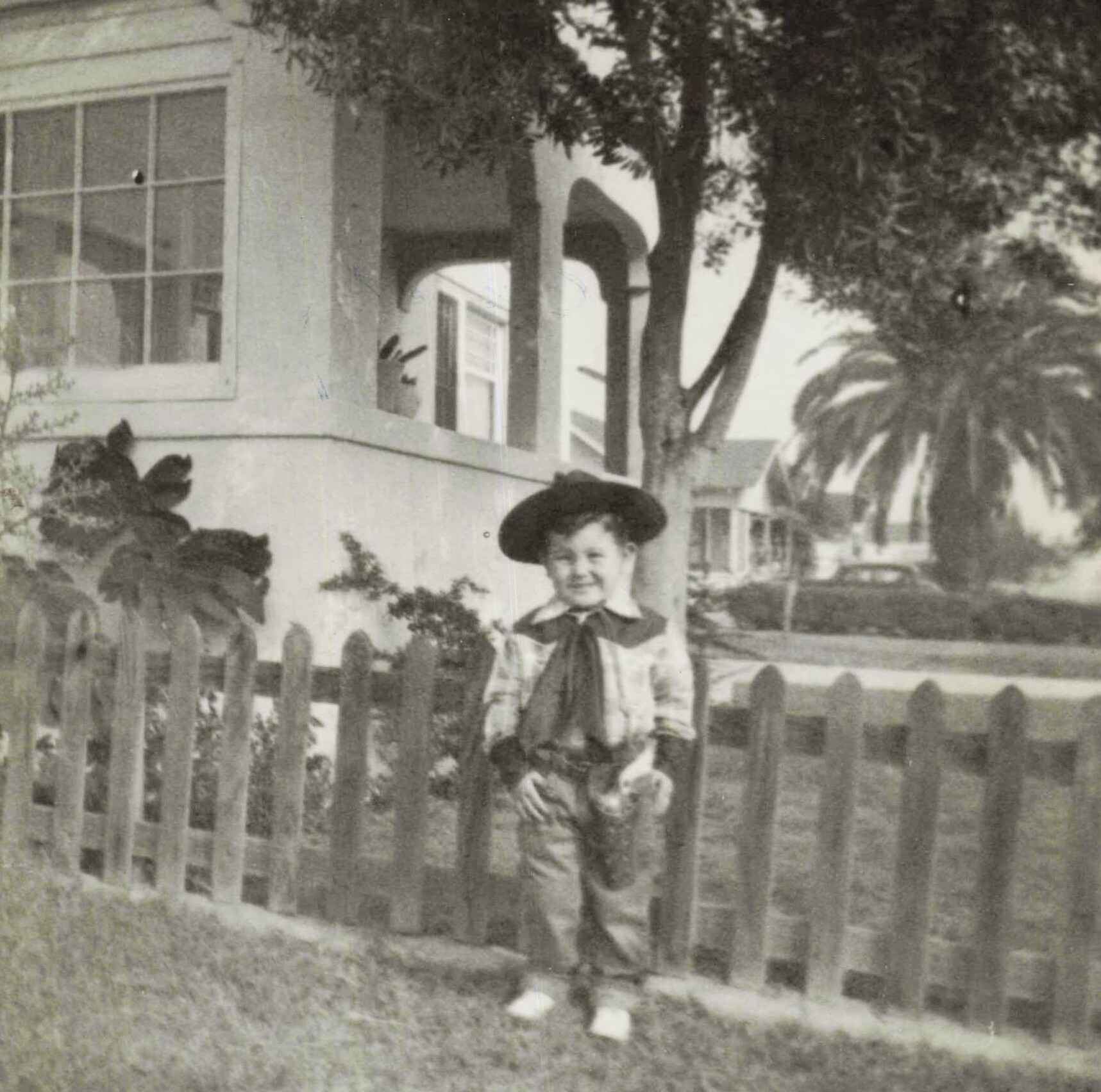 Child in a wide-brim hat standing in front of a wooden picket fence by a house with large windows, in a garden.