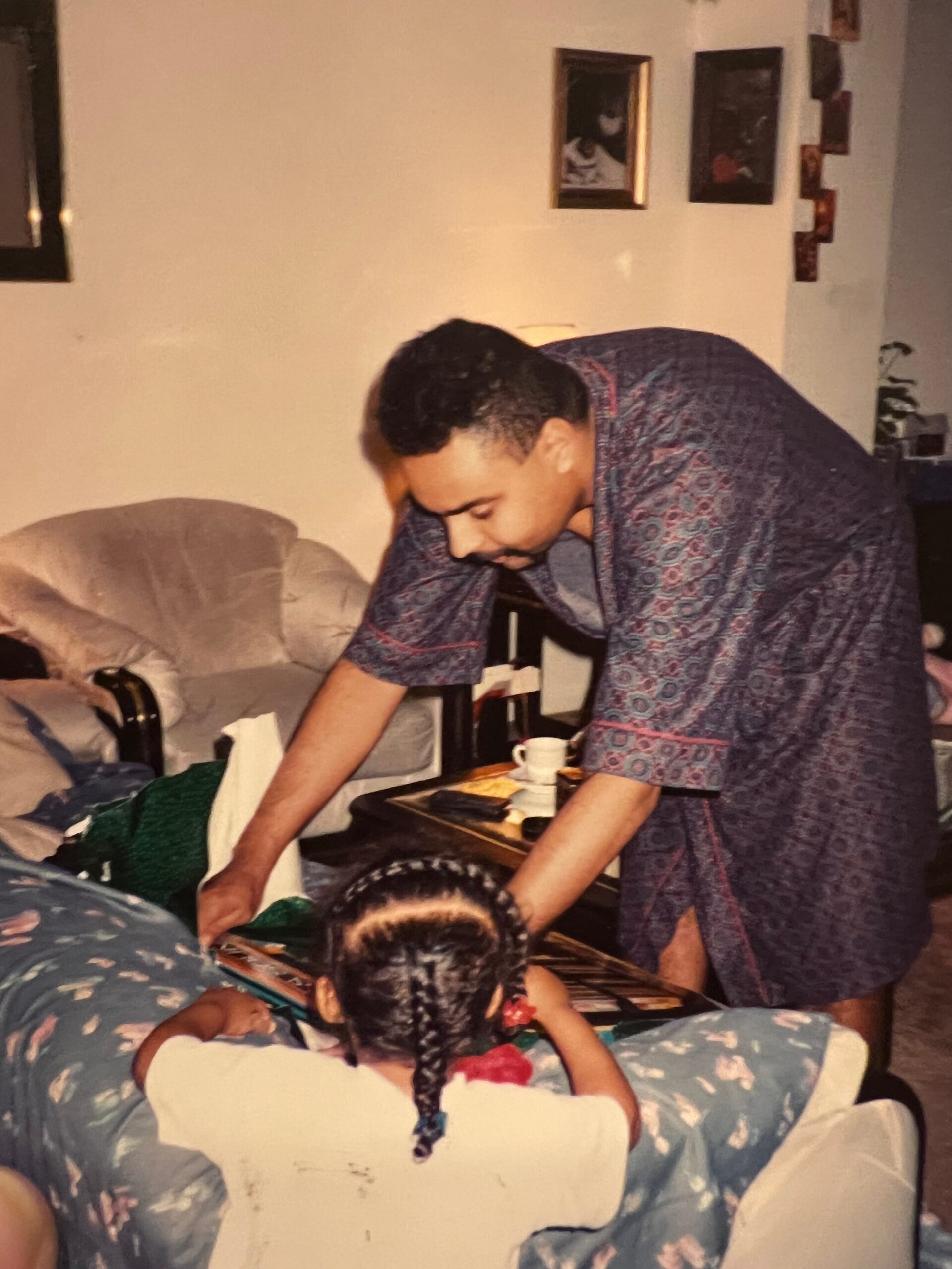 Man in a patterned robe bends over a table in a living room, with kids seated on a floral couch in the foreground.