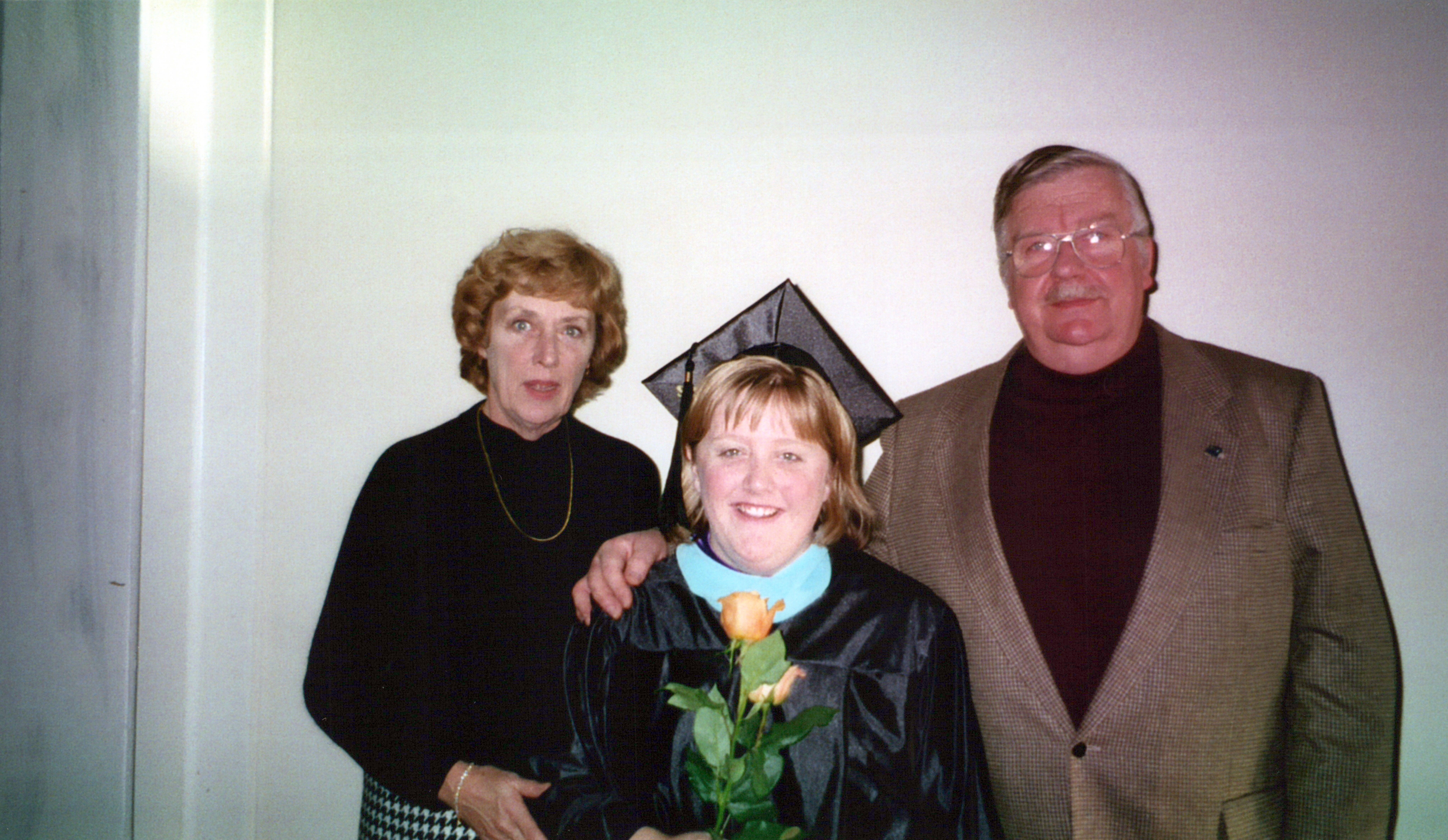 Smiling graduate in cap and gown holding yellow roses, flanked by her parents or relatives in a hallway photo.