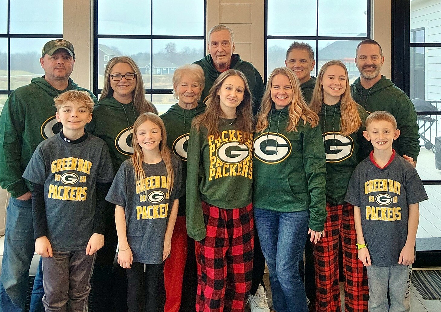 Group of people of varying ages posing together indoors, all wearing Green Bay Packers apparel (shirts and hoodies).