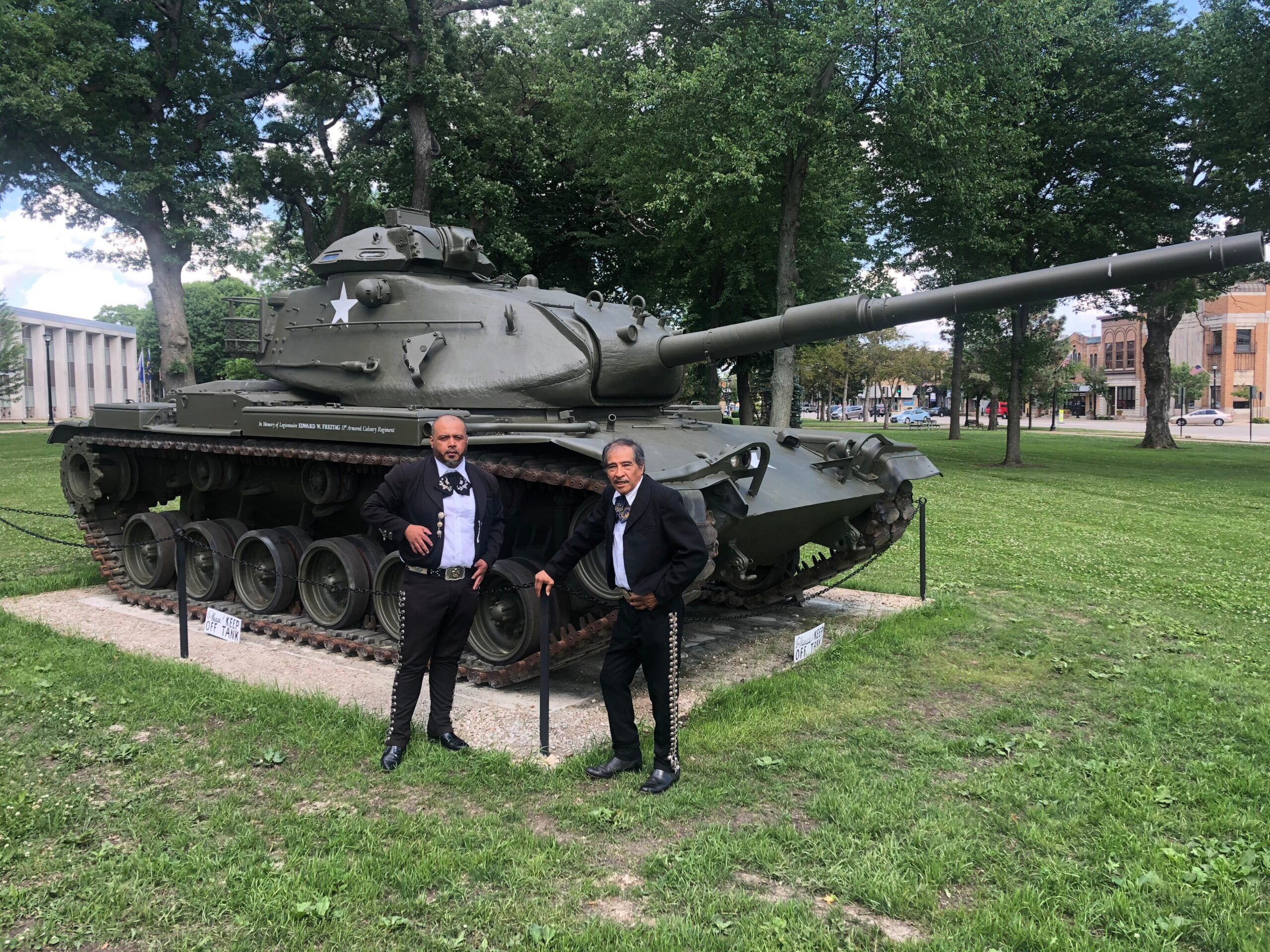 Public park display of a large military tank with a long gun barrel; two men in formal suits pose nearby under trees.