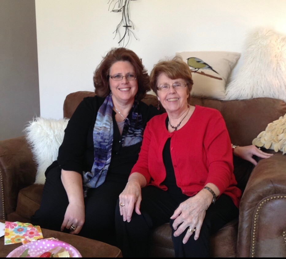 Two smiling women sit close together on a brown leather couch in a living room, one wearing black with a blue scarf and glasses, the other in a red cardigan with glasses and jewelry.