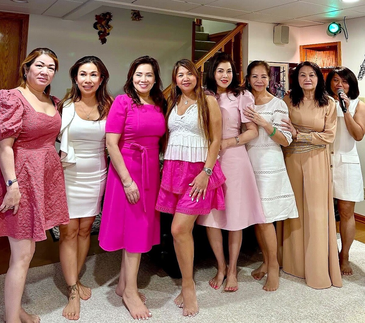 Group of eight women in pink and white dresses posing together indoors; one woman on the right speaks into a microphone.