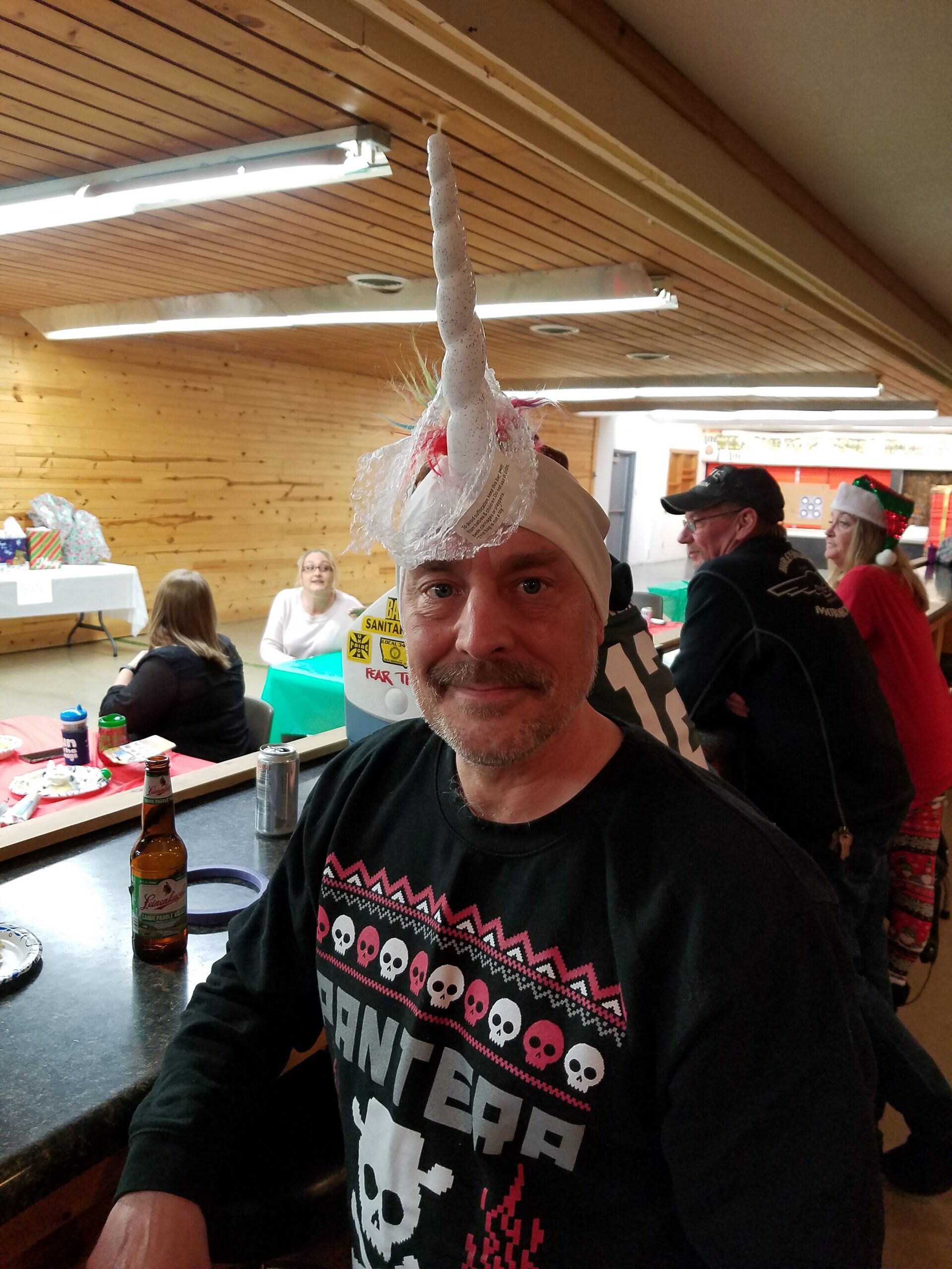 Man wearing a white unicorn-horn headpiece at a festive indoor party, with drinks and presents on tables in the background