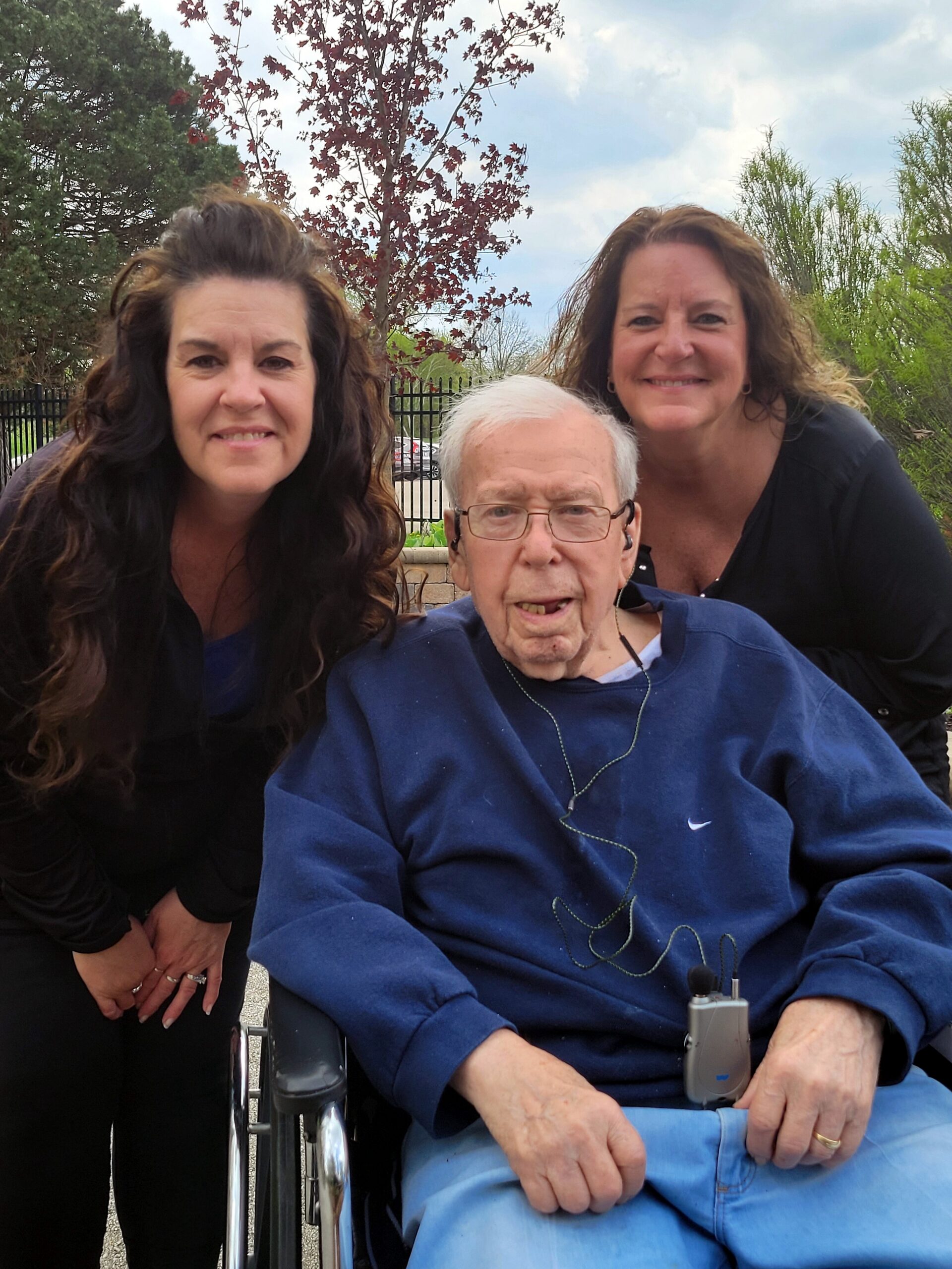 Three smiling people outdoors: two women stand behind an elderly man in a wheelchair, on a sunny day with trees and a fence in the background.