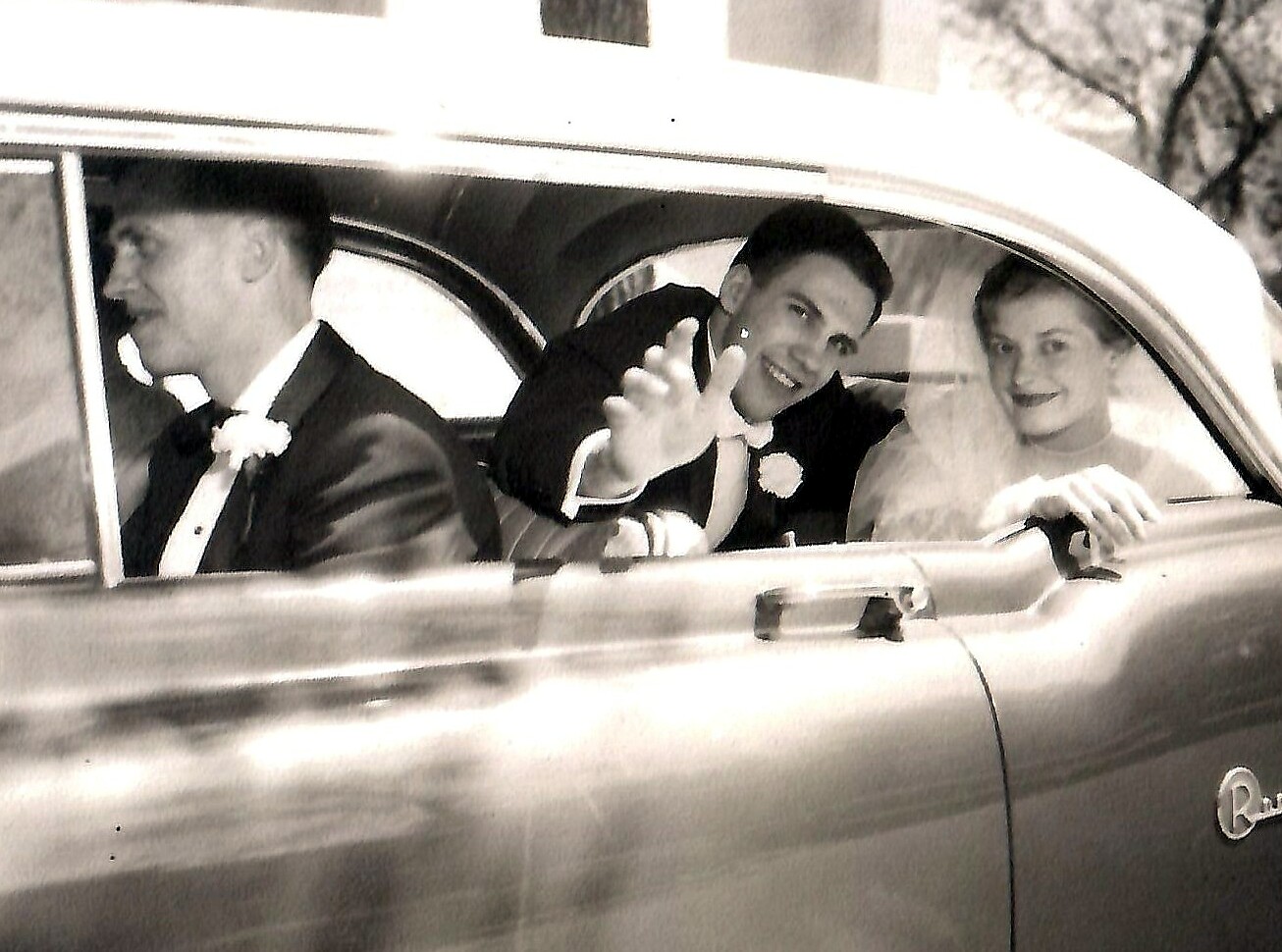 Bride and groom ride in a vintage convertible, the groom in the back waves to the camera while the bride smiles in the front seat; driver in tuxedo on left side.