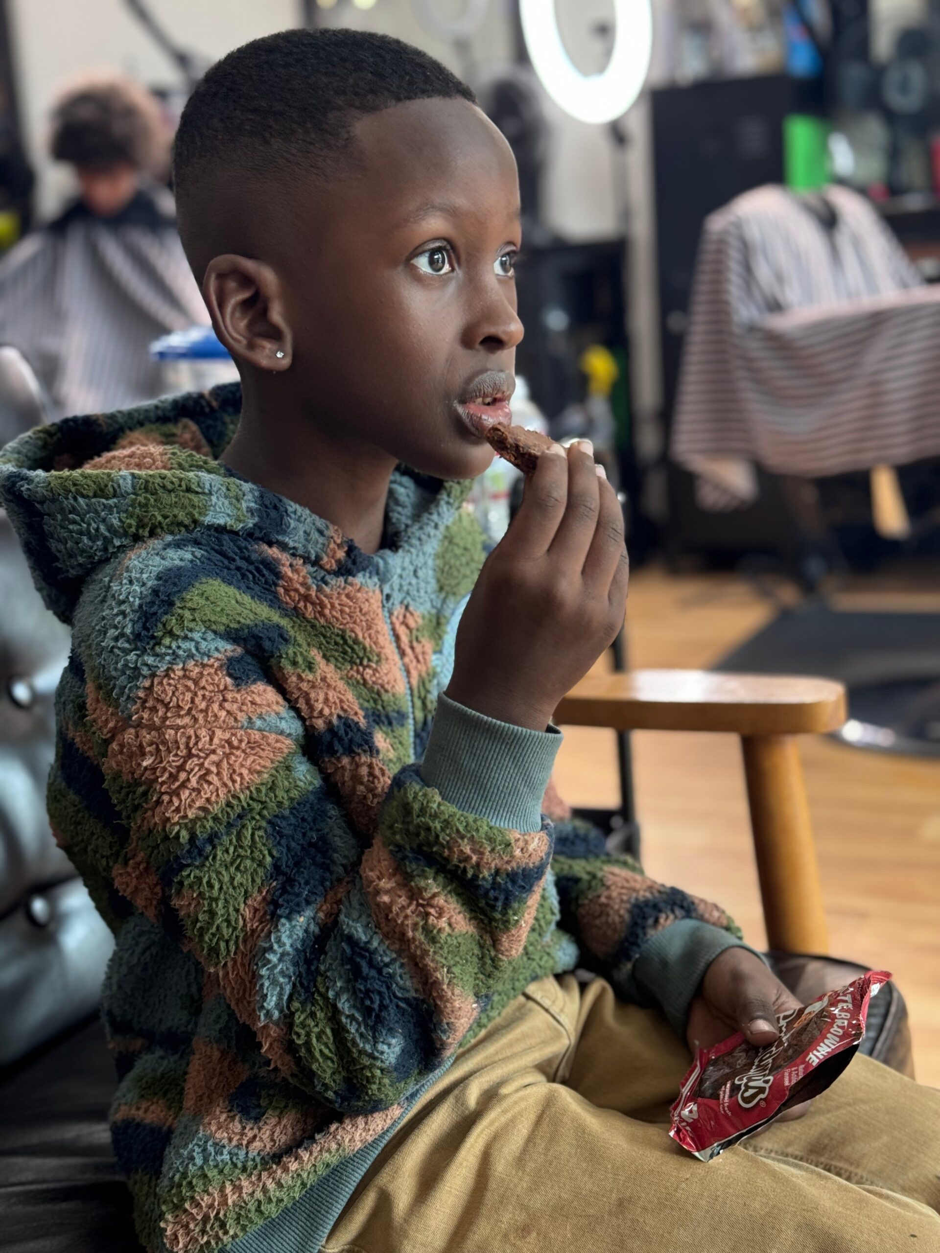 Boy in a colorful fleece hoodie eats a snack while seated in a barbershop.