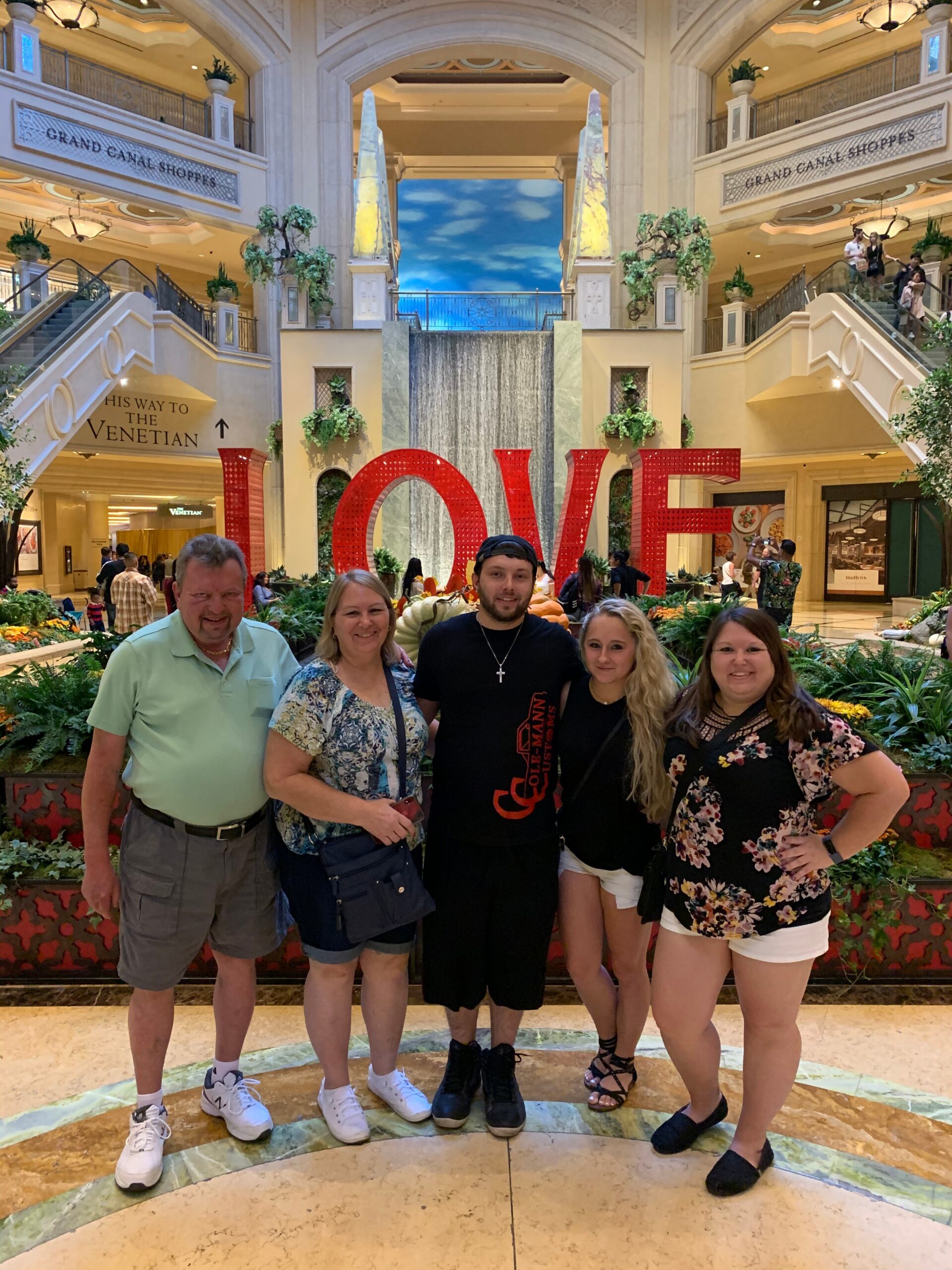 Five people pose in front of a large red LOVE sculpture in a mall atrium with balconies, greenery, and a waterfall backdrop.