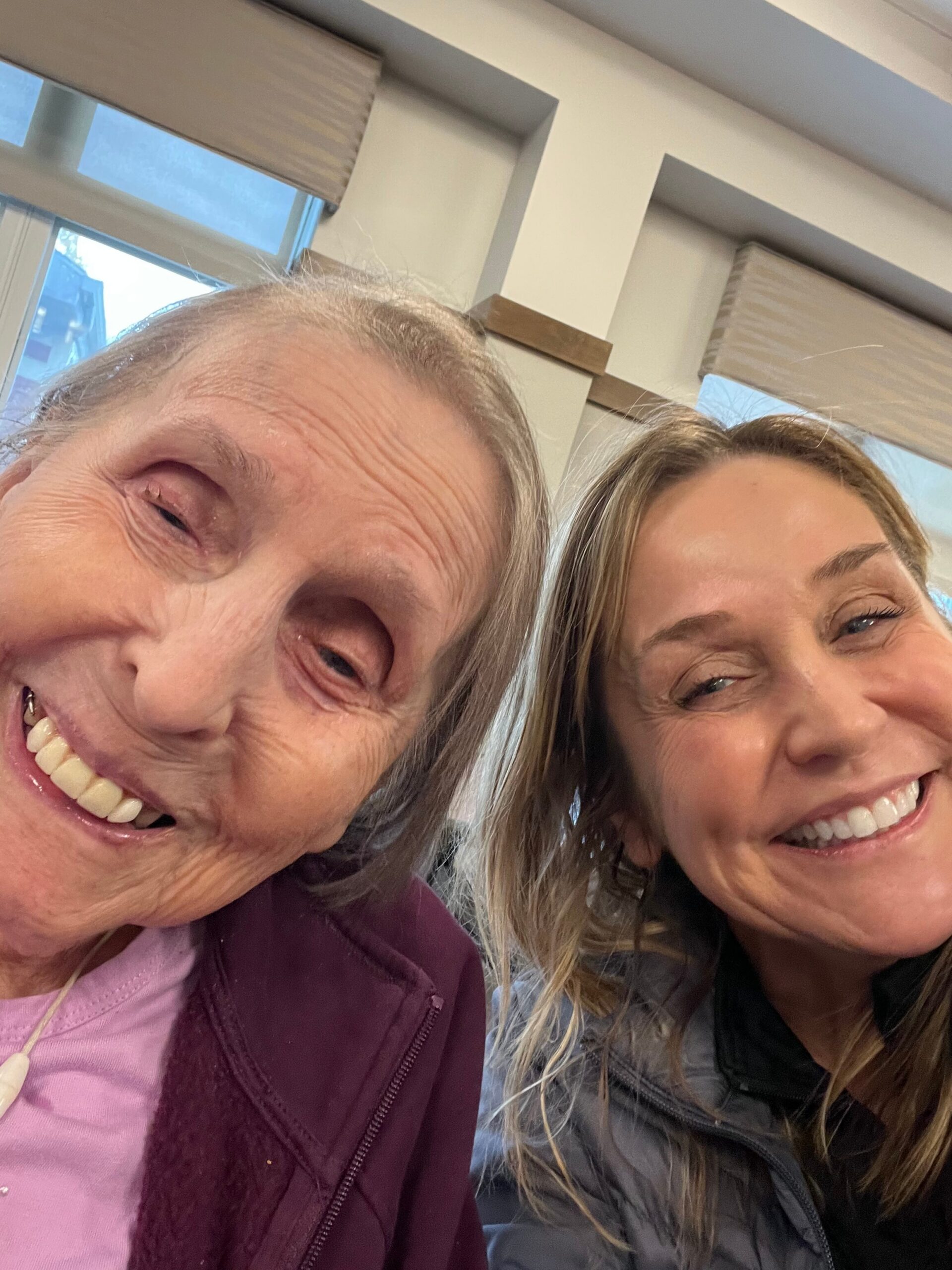 Two women smiling at the camera in a close-up selfie, one wearing a pink top and maroon jacket, the other in a dark jacket.