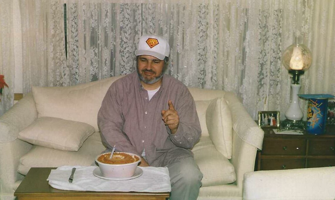 Man on a beige sofa wearing a white cap, pointing at the camera, with a bowl of food on the coffee table in front.