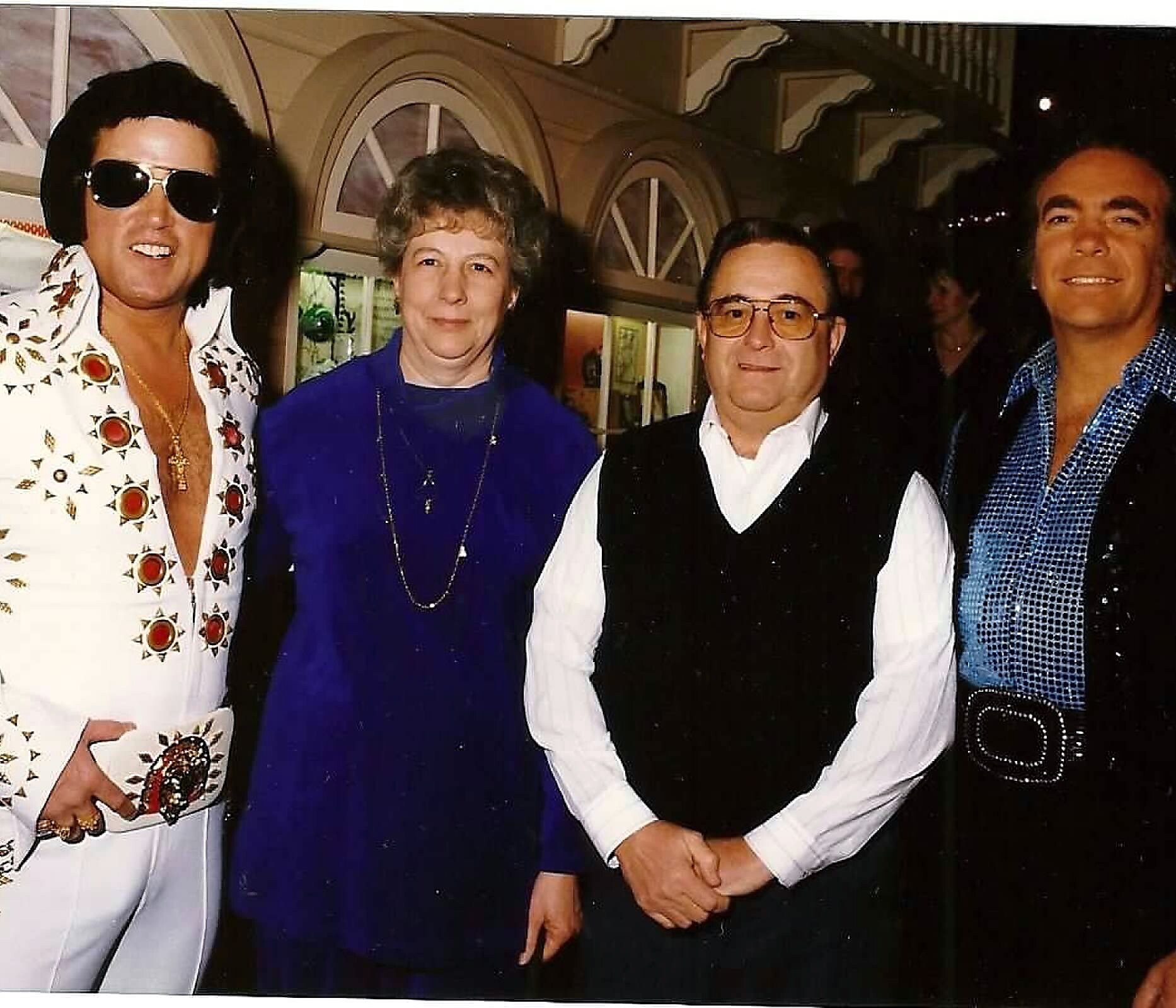 Four adults posing for a group photo at night: left man in a white patterned shirt and sunglasses, woman in blue, man with glasses in a white shirt and black vest, man in a blue checked shirt on the right, arched windows in the background.