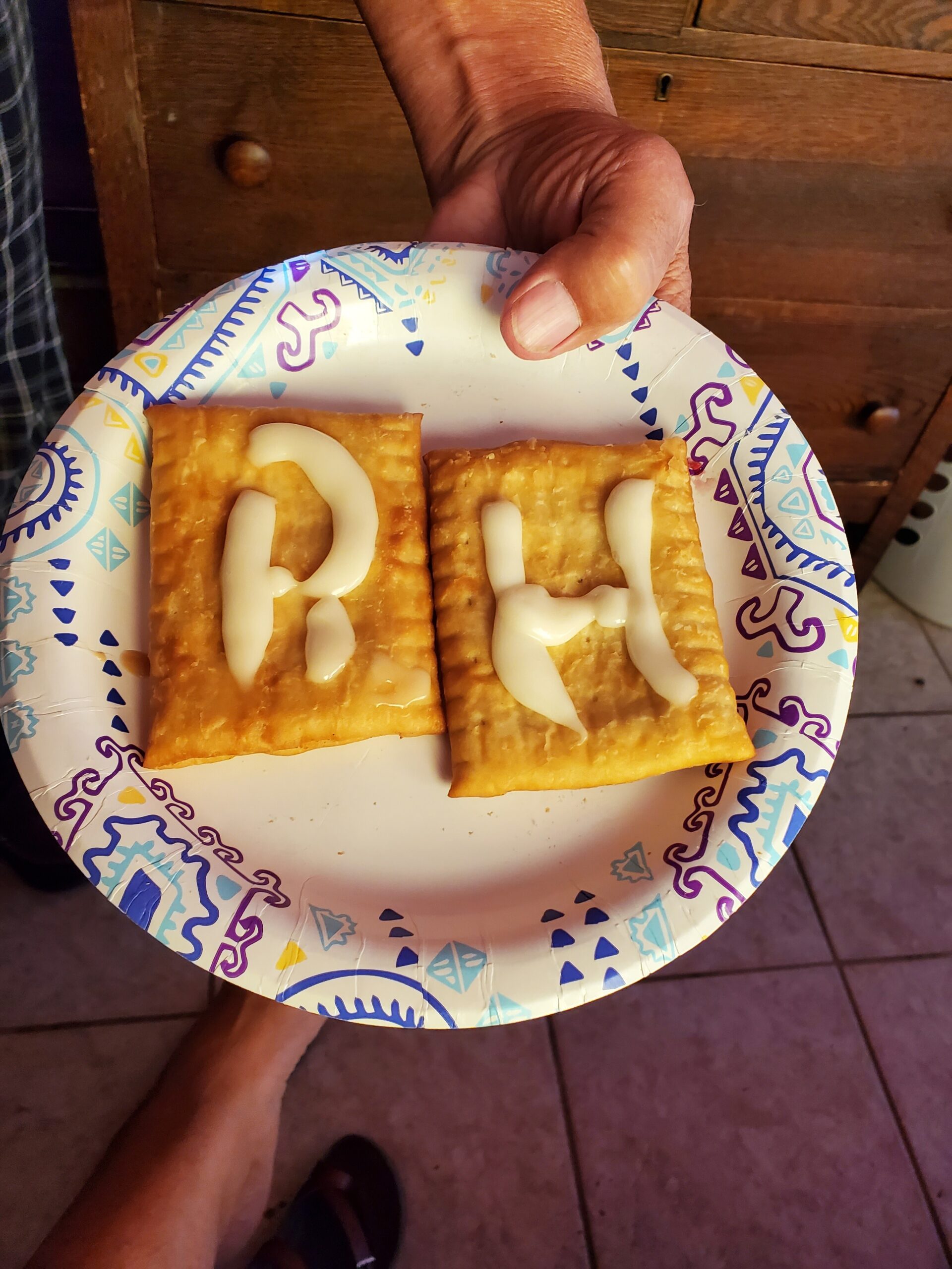 Two square crackers with white icing forming letters on a colorful patterned plate, held by a person's hand.