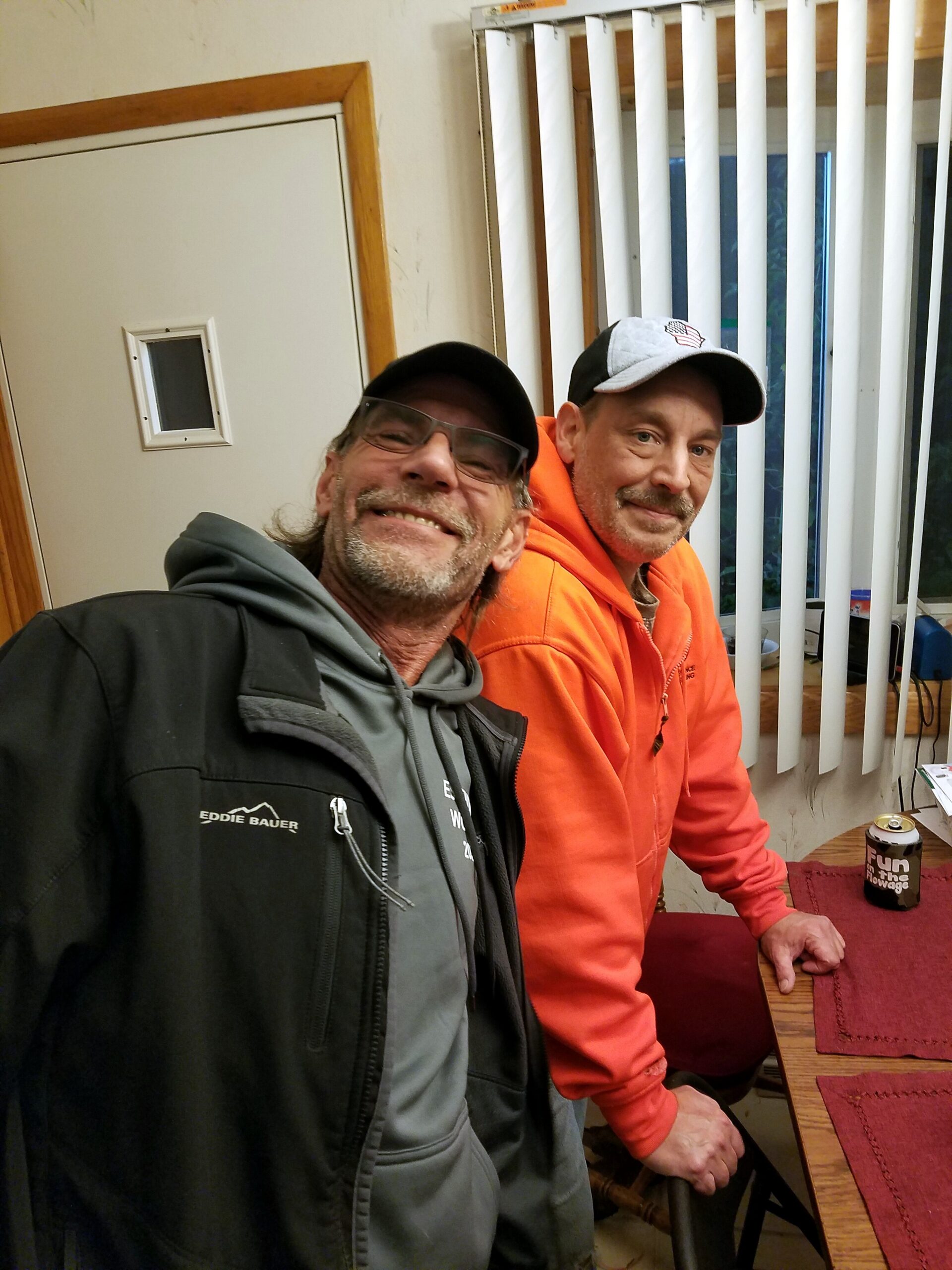 Two men smile at the camera in a cluttered room, one in a gray hoodie and black jacket, the other in an orange hoodie and cap.