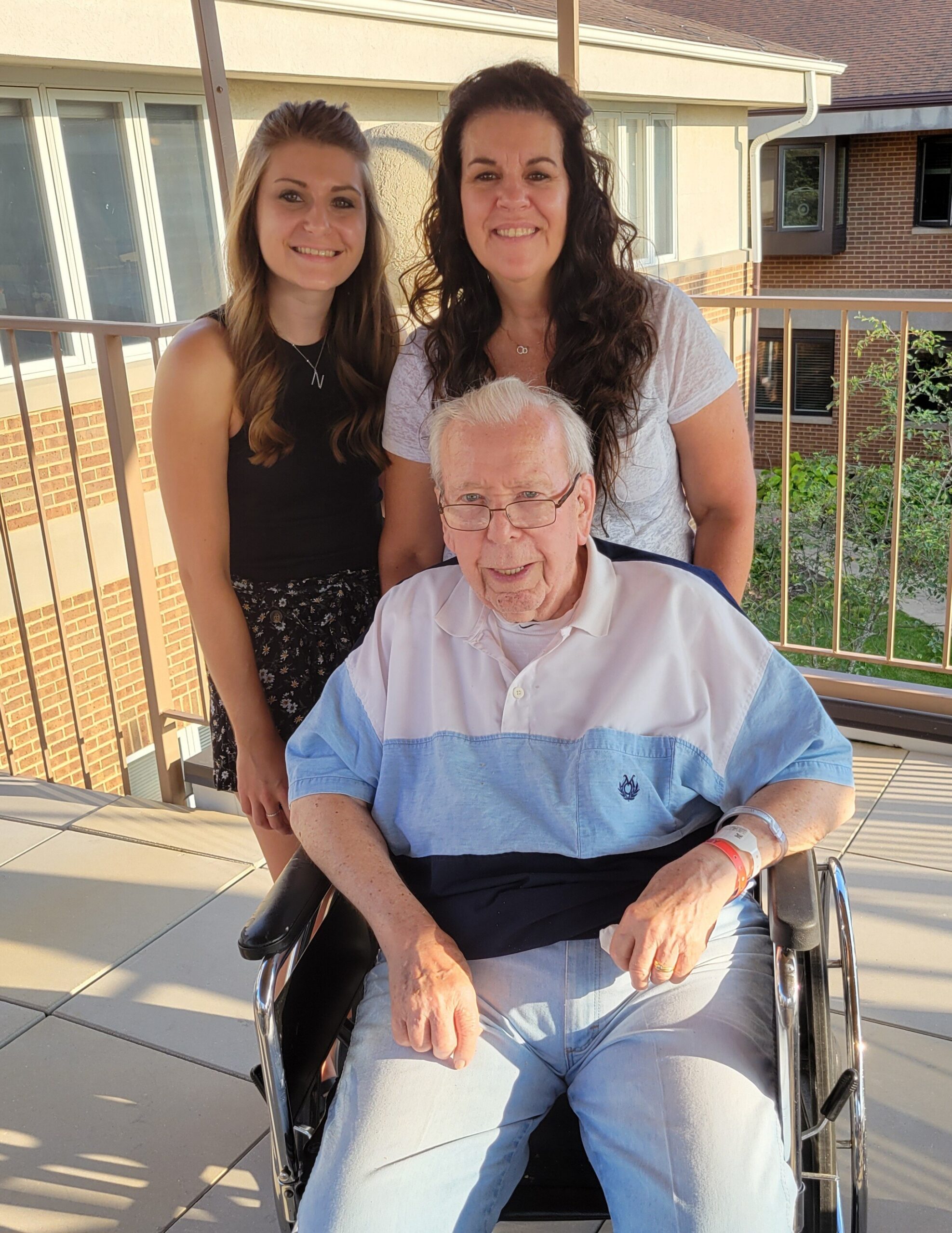Elderly man in a wheelchair on a balcony, flanked by two smiling women.