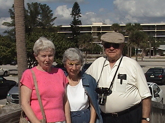 Three elderly friends posing in a sunny parking lot; two women in pink and white outfits and a man with a hat and a camera hanging from his neck.