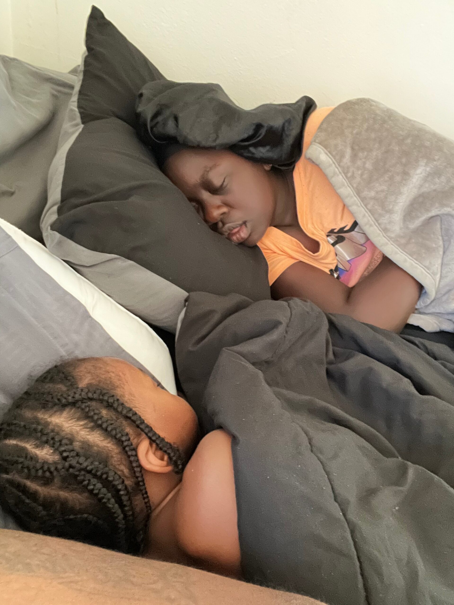 Young child asleep on a bed wearing an orange shirt, covered by a gray comforter with gray pillows nearby.