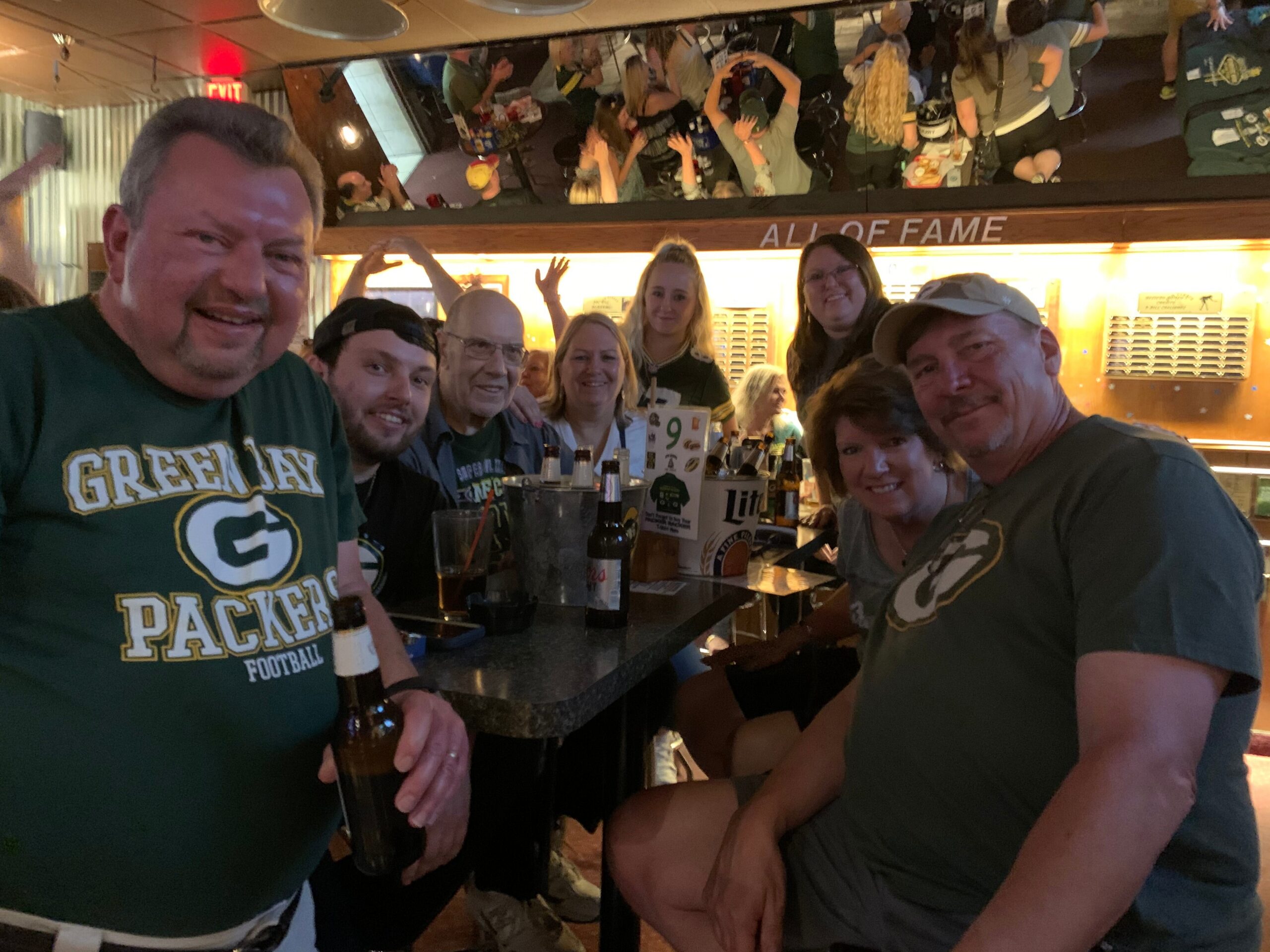 Group of Packers fans posing at a bar, wearing green jerseys, smiling with drinks for a selfie-style photo outside the frame.