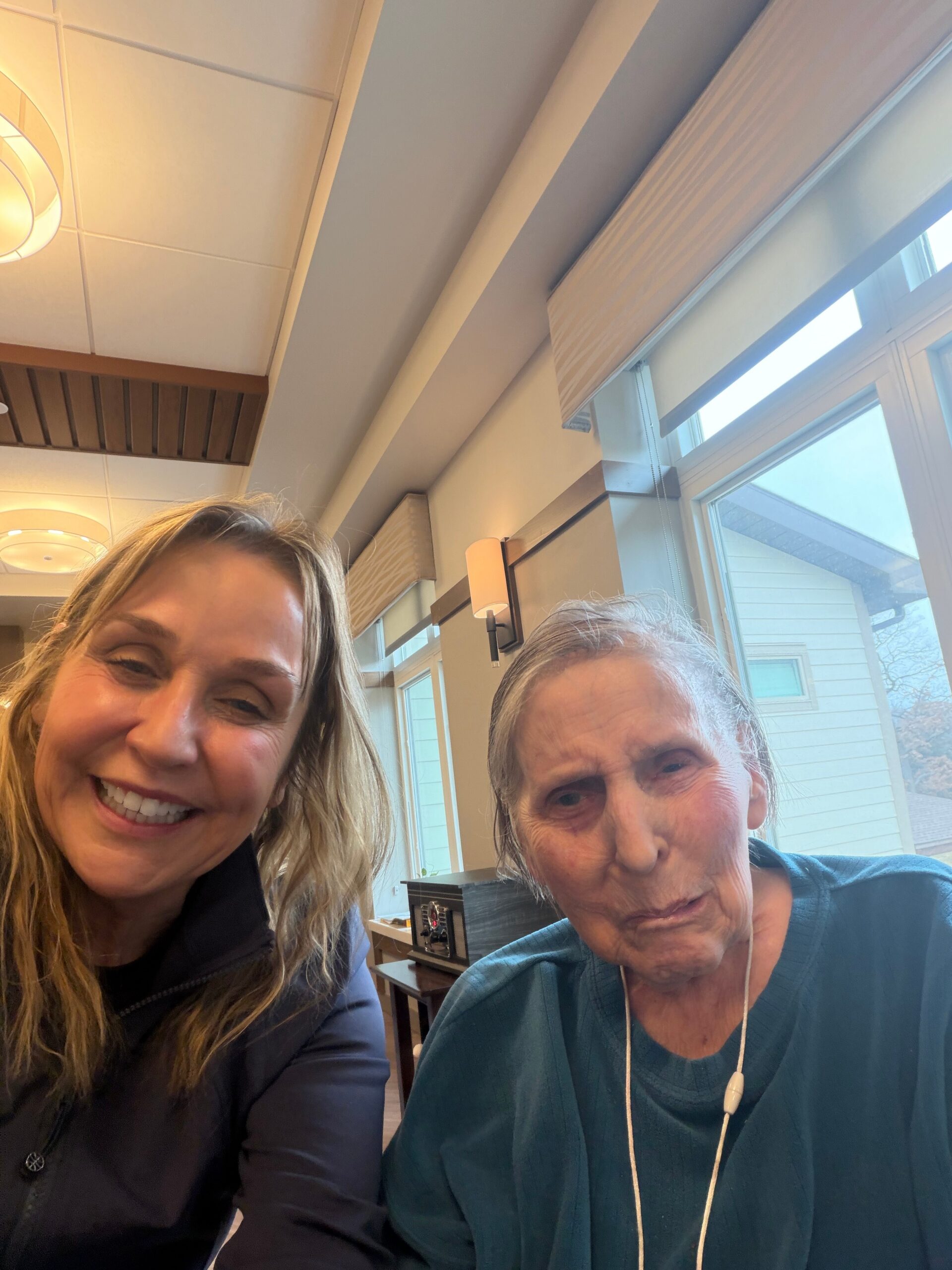 Two women are taking a cheerful selfie indoors, with one younger woman on the left and an older woman on the right near large windows with blinds.