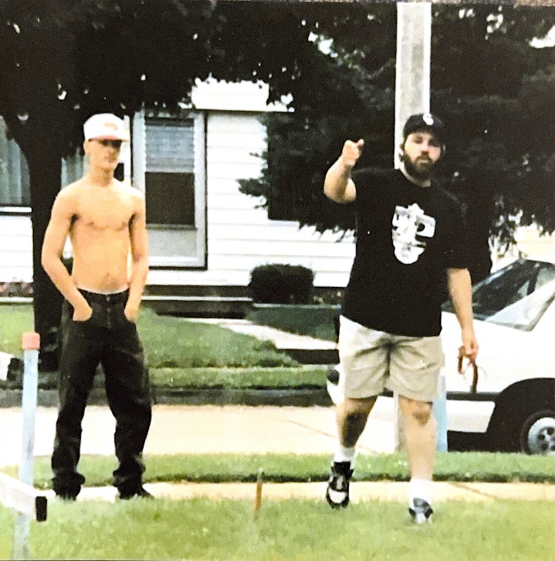 Two men standing on a manicured lawn; one shirtless in a cap, the other with a beard and a prosthetic leg, pointing at the camera.