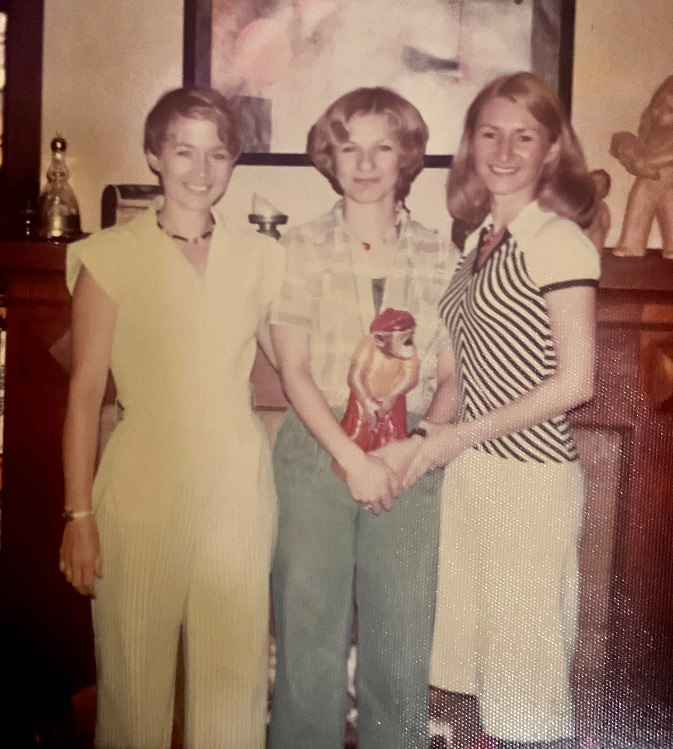 Three adults posing together indoors, smiling for the photo. A woman in yellow on the left, a person with light hair in the middle, and a woman in a striped dress on the right.