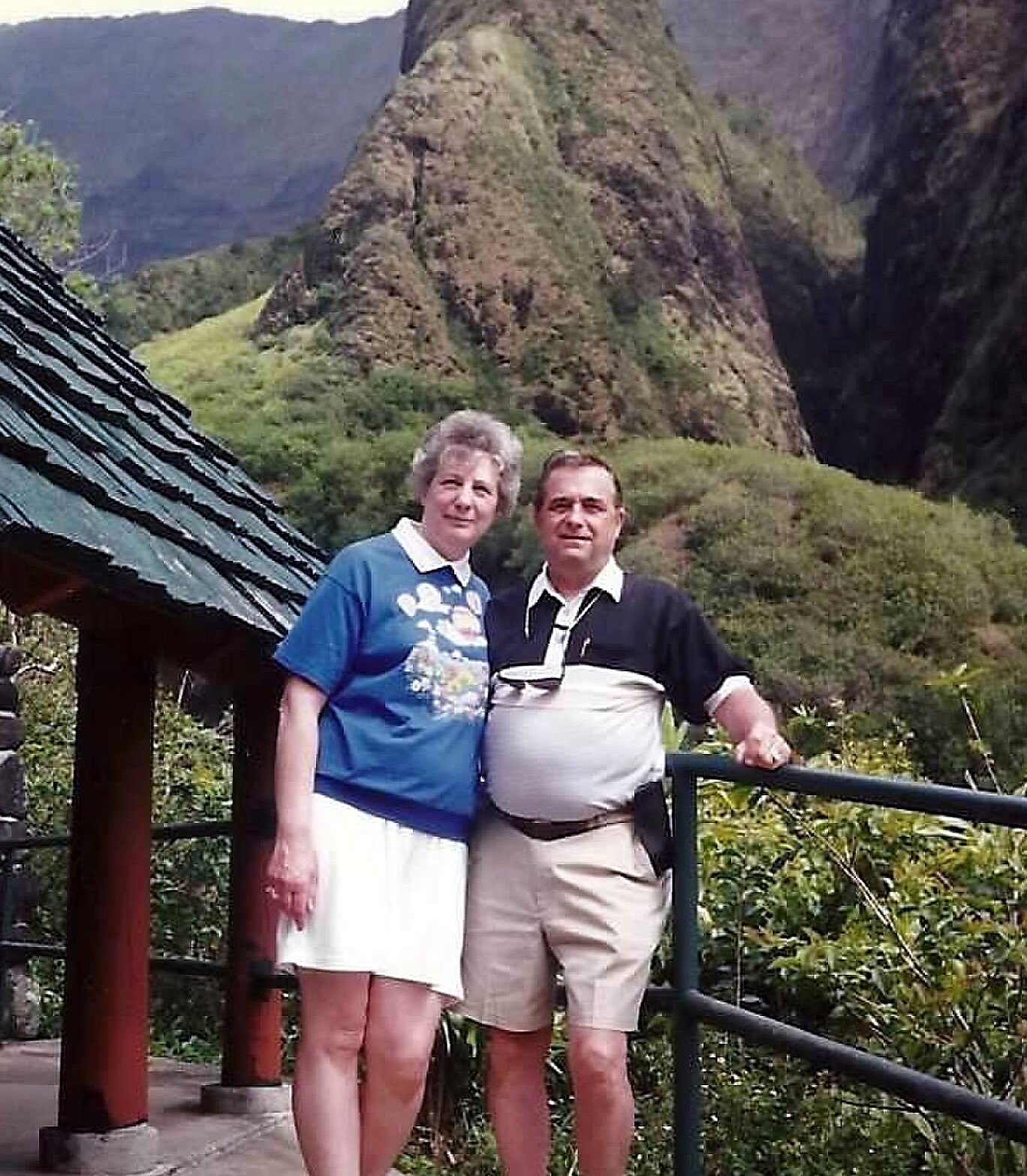 Two people posing together on an overlook with rocky mountains behind them, under a bright sky.