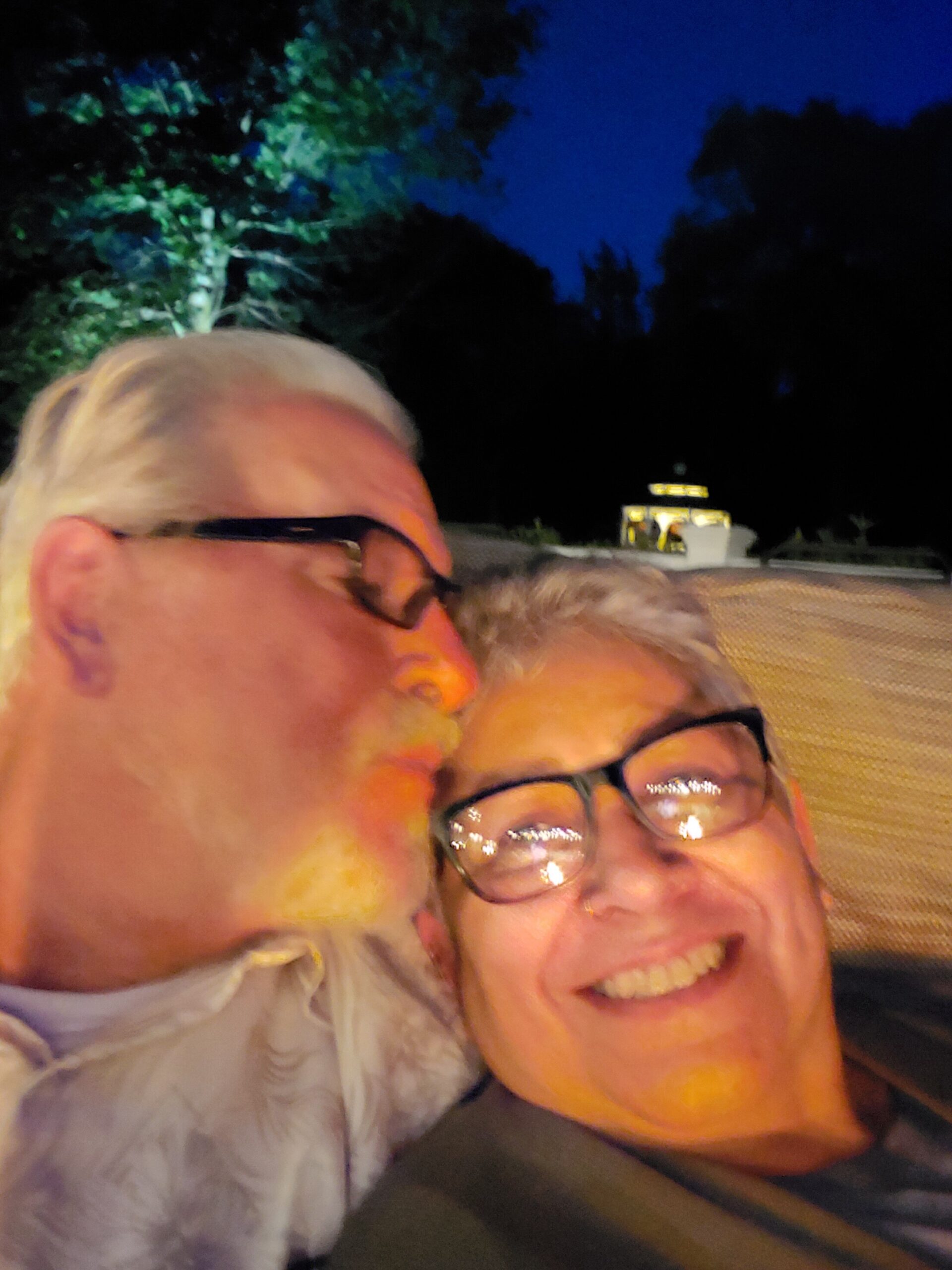 Two people smiling for a close-up selfie at night outdoors, with colorful face paint on their cheeks and glasses visible.
