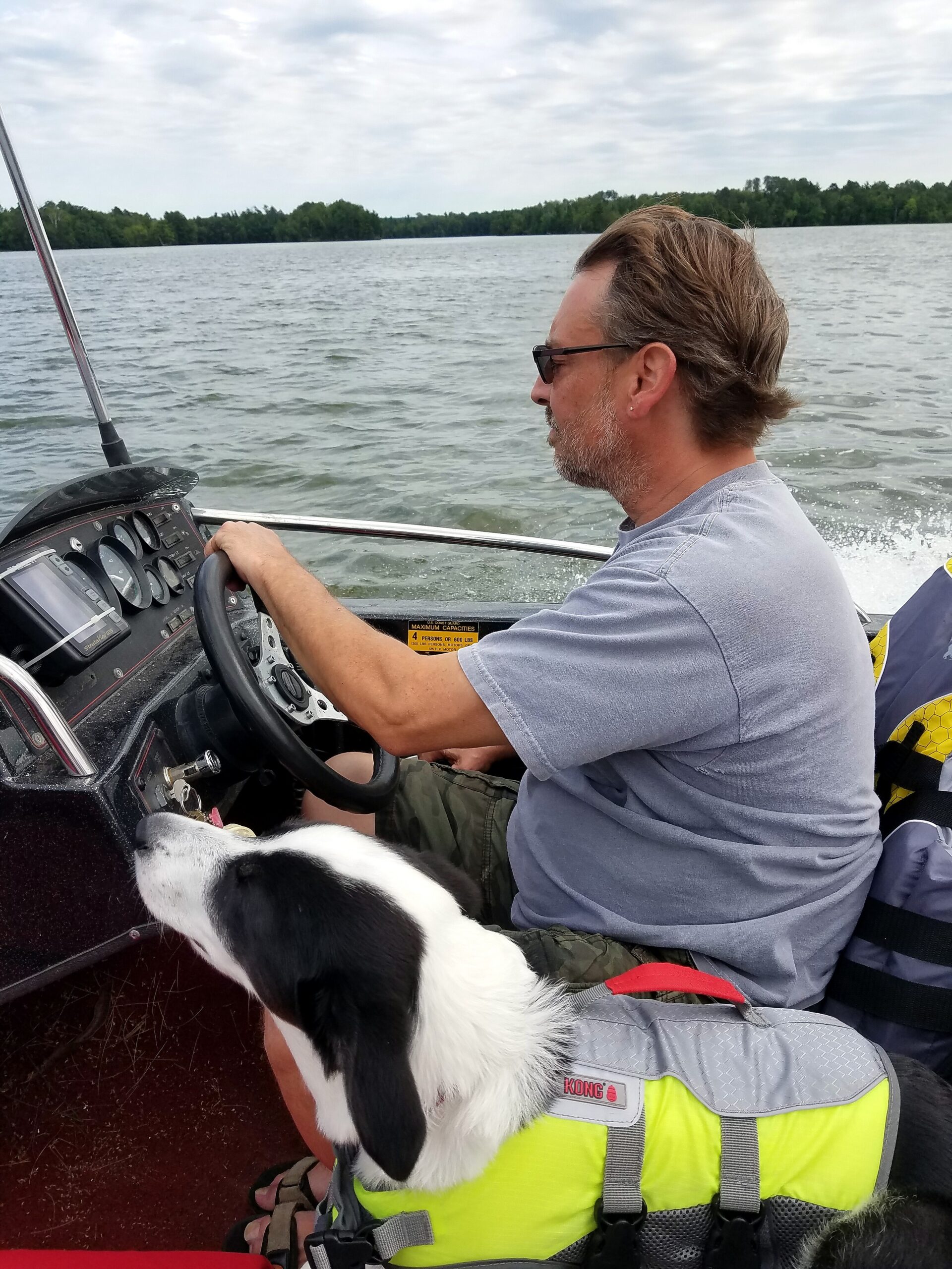 Man steering a small boat on a lake, wearing sunglasses, with a black-and-white dog sitting near the passenger seat in the foreground.