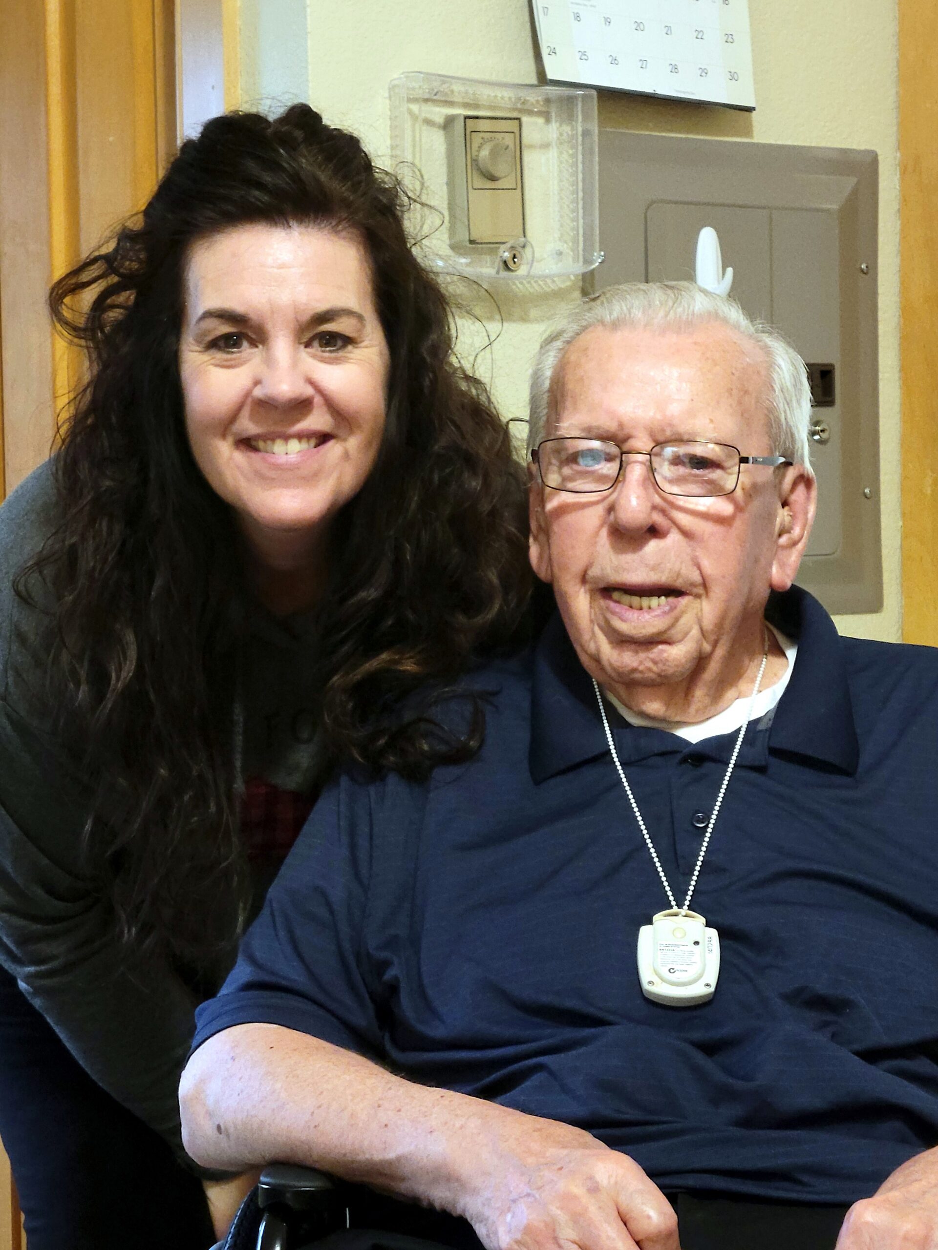 Smiling woman with long dark hair sits beside an elderly man wearing a navy polo and a medical alert pendant on a lanyard, both in a doorway setting (likely a care facility).