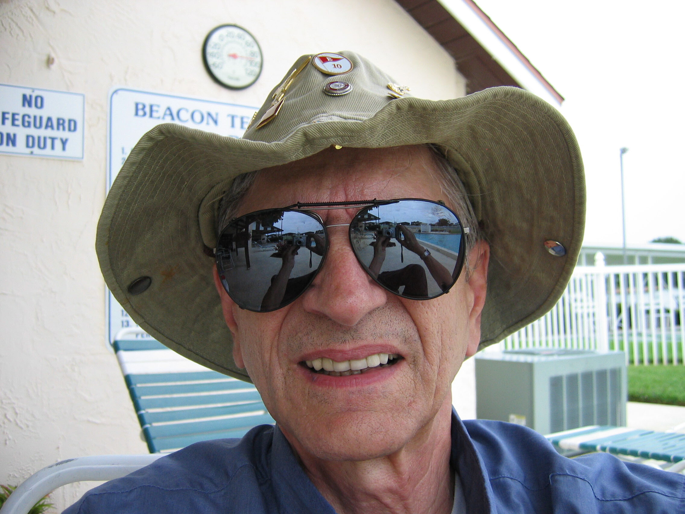 Close-up of a man wearing a wide-brimmed hat and mirrored sunglasses, with a poolside reflection visible in the lenses.