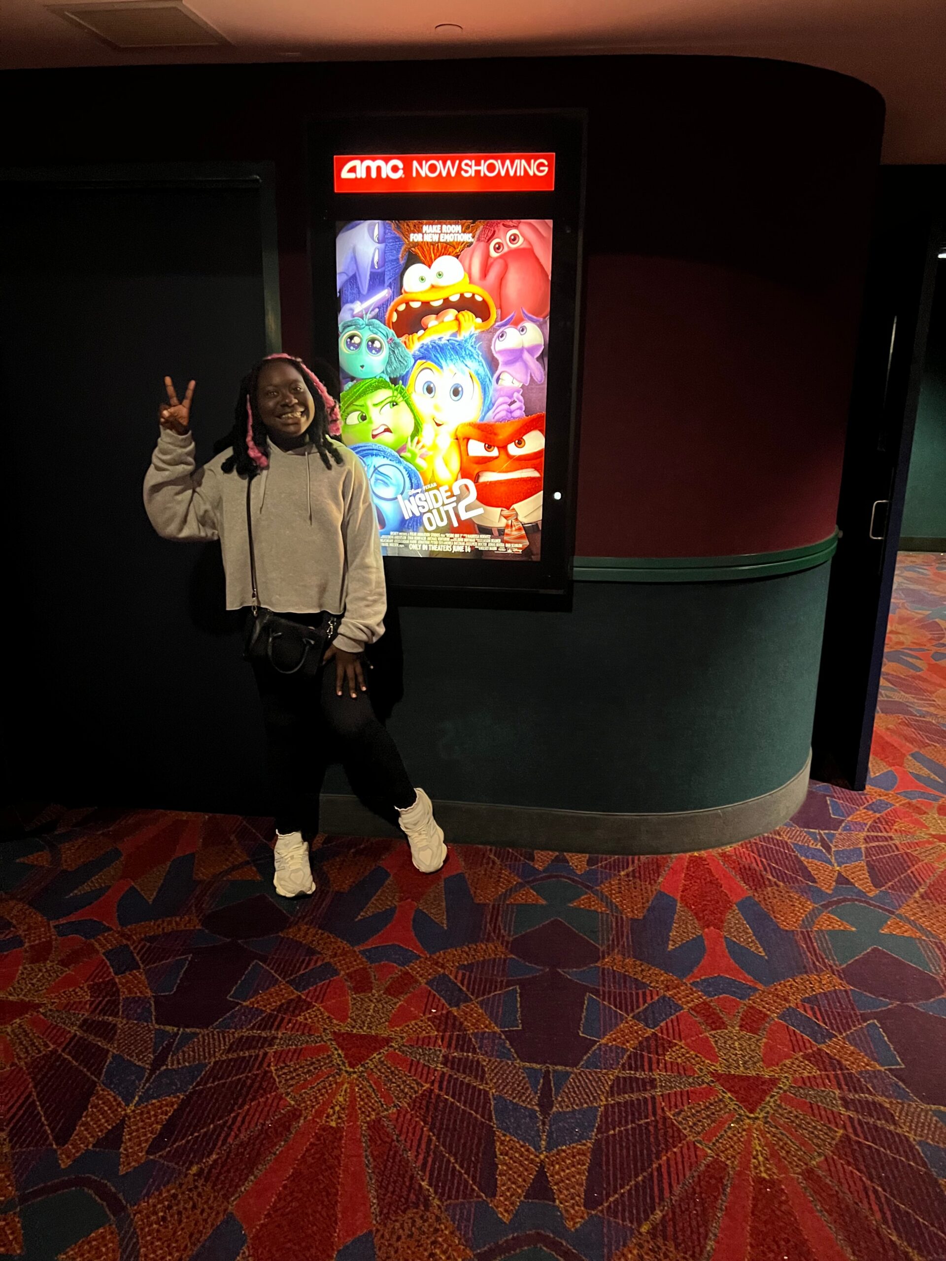 Person posing with a bright Inside Out 2 movie poster at an AMC Now Showing stand, making a peace sign with one hand.