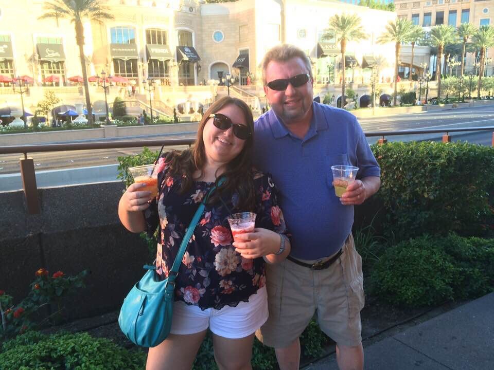 Two smiling adults wearing sunglasses, holding drinks while posing outdoors in a sunny city setting with storefronts in the background.