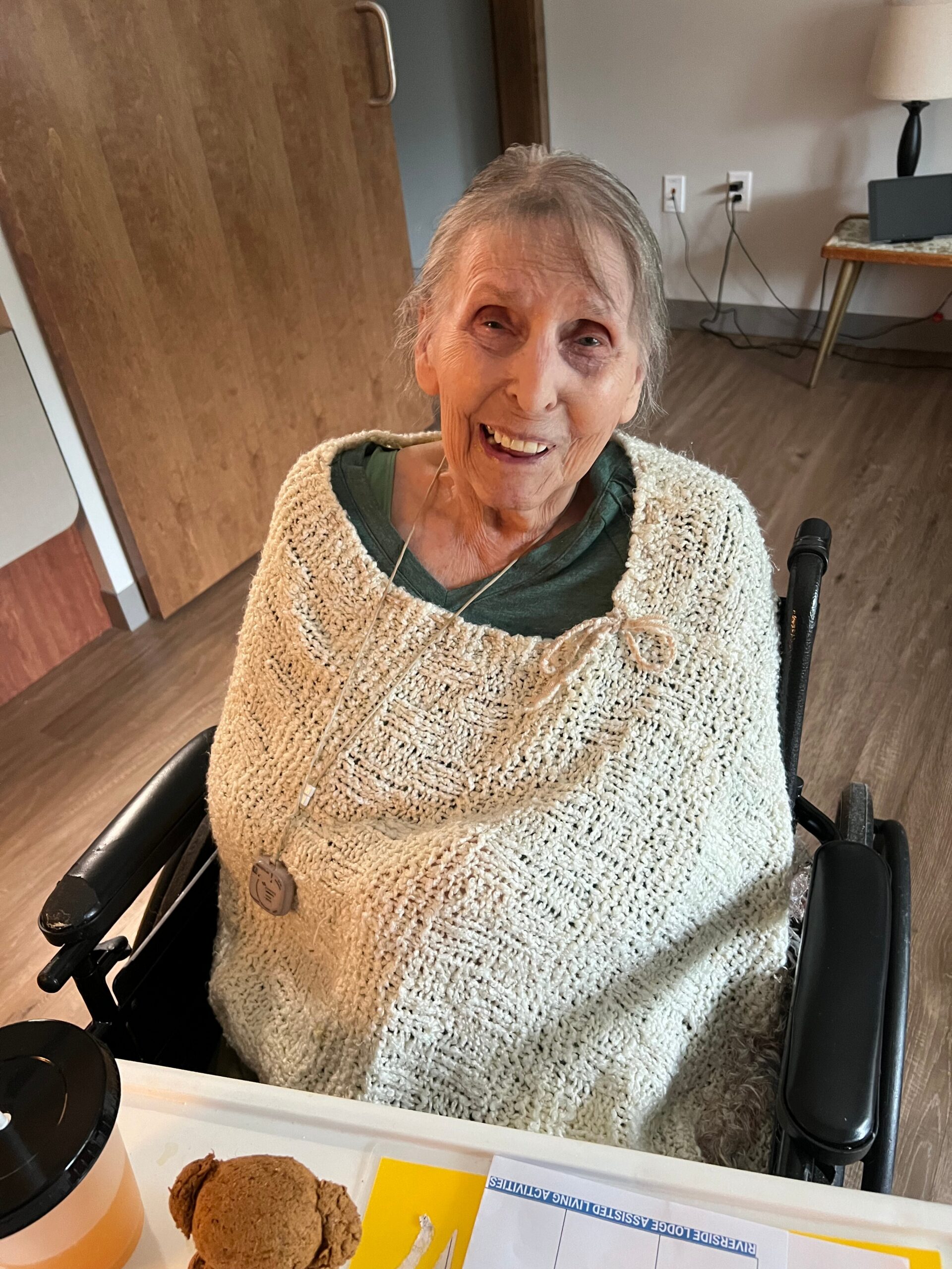 Smiling elderly woman in a wheelchair wearing a cream knitted shawl, seated at a table with cookies and coffee nearby.