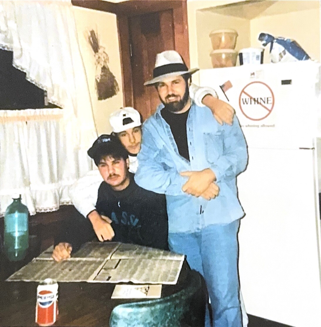 Three men posing in a cluttered kitchen; one seated with a newspaper, two standing (one with a hat); fridge in the background with a 'WHINE' sign.