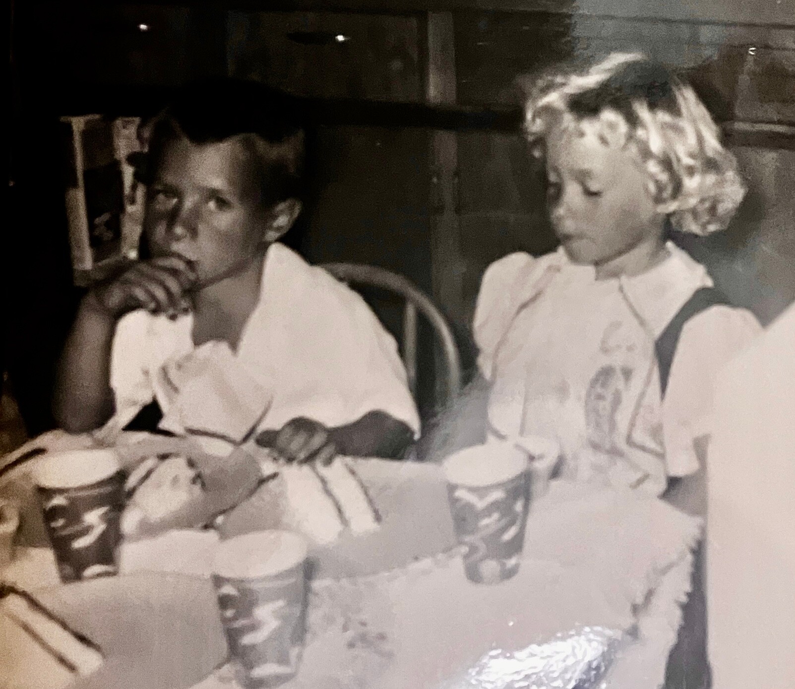 Two children sit at a table eating, with paper cups and plates in front of them; the boy on the left rests his chin on his hand as he looks ahead.