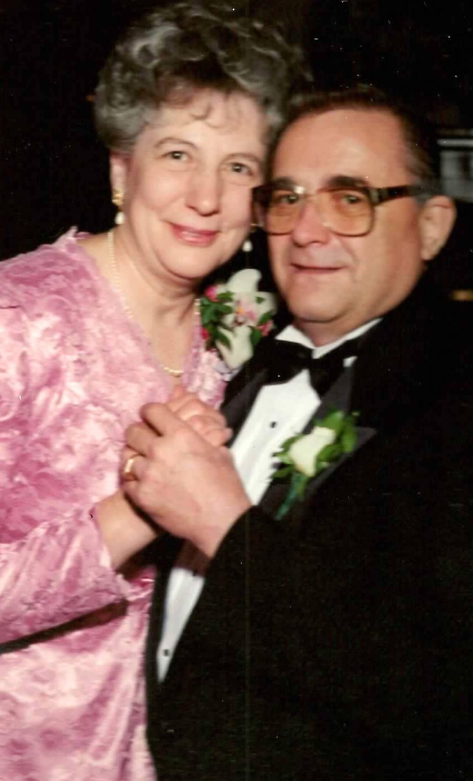 Older couple dancing at a formal event; woman in pink lace dress with pearls, man in a tuxedo and glasses, smiling at the camera.
