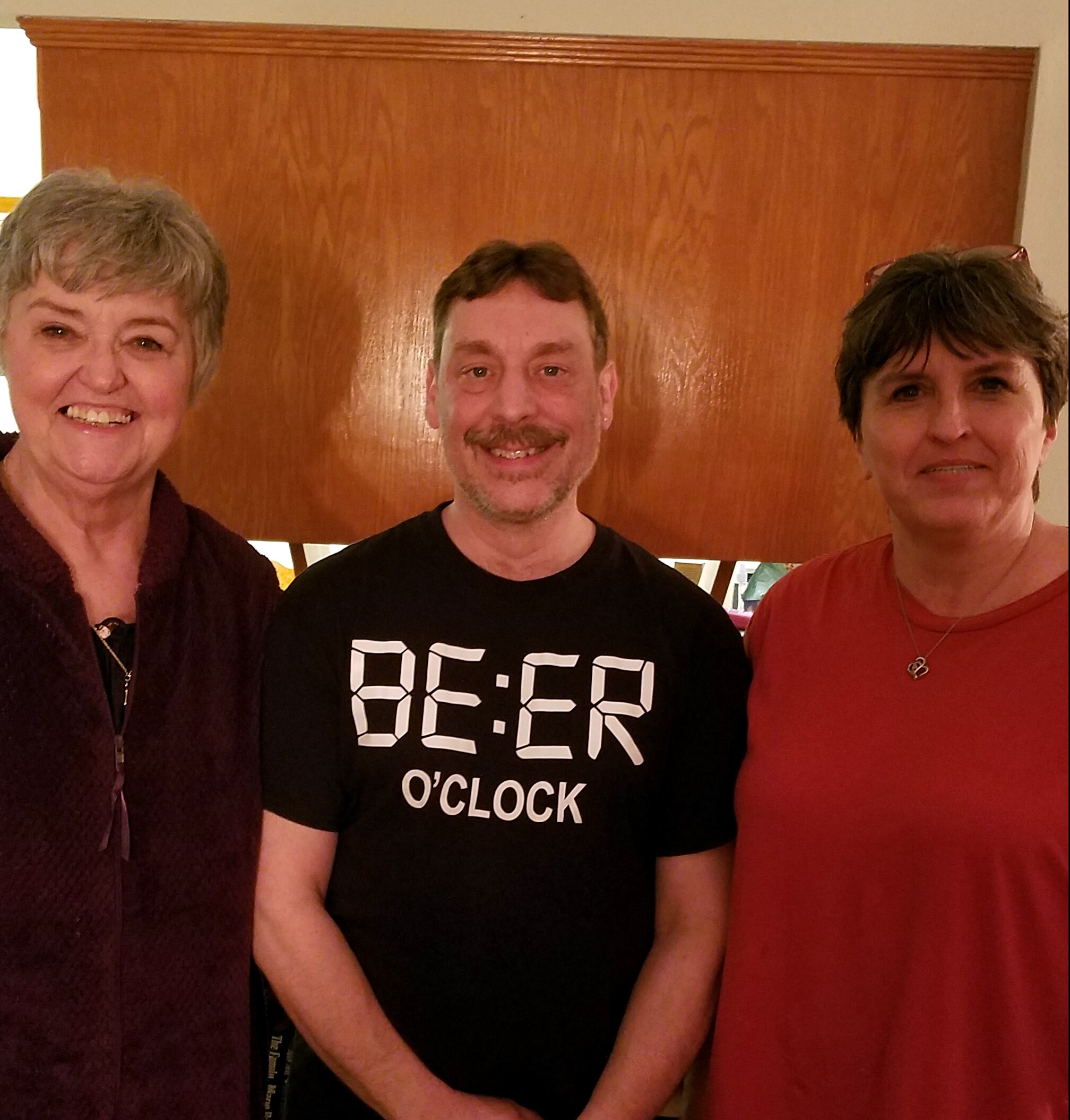 Three smiling adults pose together indoors in front of a wooden door; left woman with short gray hair, center man with a beard in a black shirt, right woman in red top.
