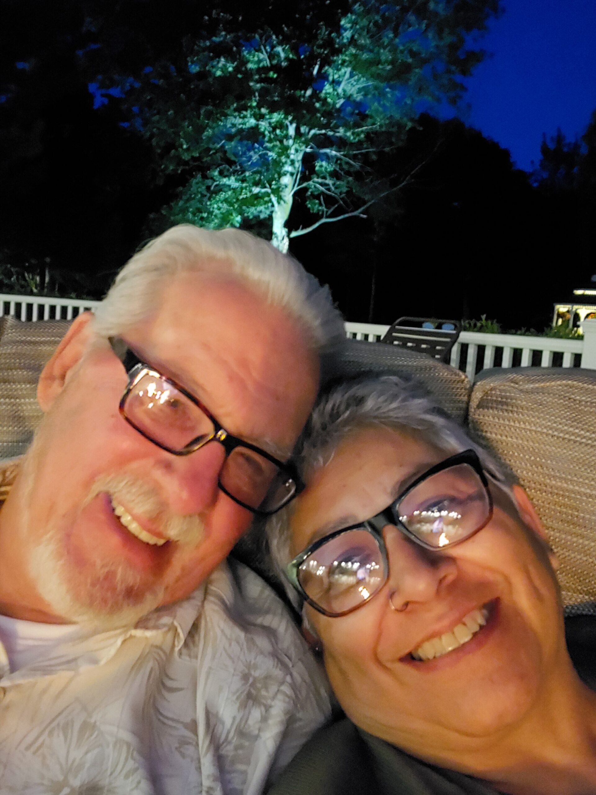 Close-up selfie of an older couple smiling at night on a patio, with a tree and outdoor furniture in the background.