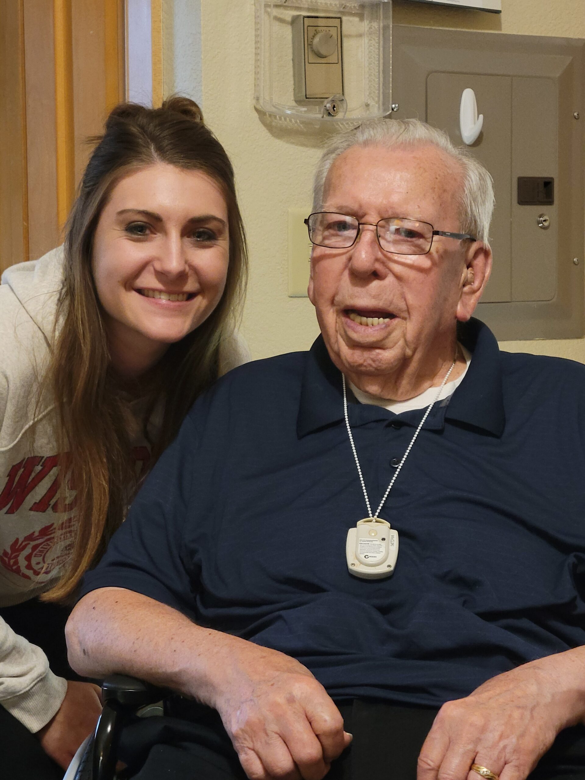 Young woman and elderly man smiling together in a care setting; the man wears glasses and a medical alert pendant on a chain.