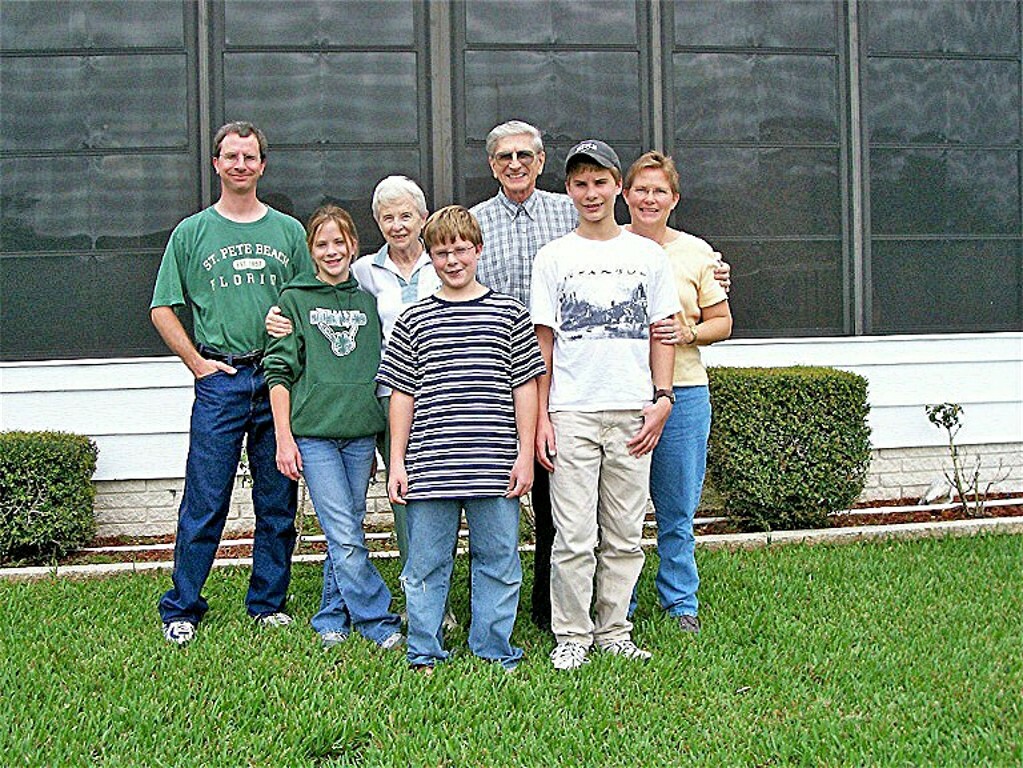 Group of seven people posing outside on a grassy lawn in front of a building, smiling at the camera.