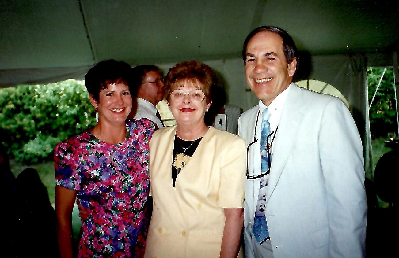Three smiling adults posing together under a tent at an outdoor event: a floral-dressed woman on the left, a woman in a pale yellow blazer in the center, and a man in a light suit on the right with glasses hanging from his shirt.