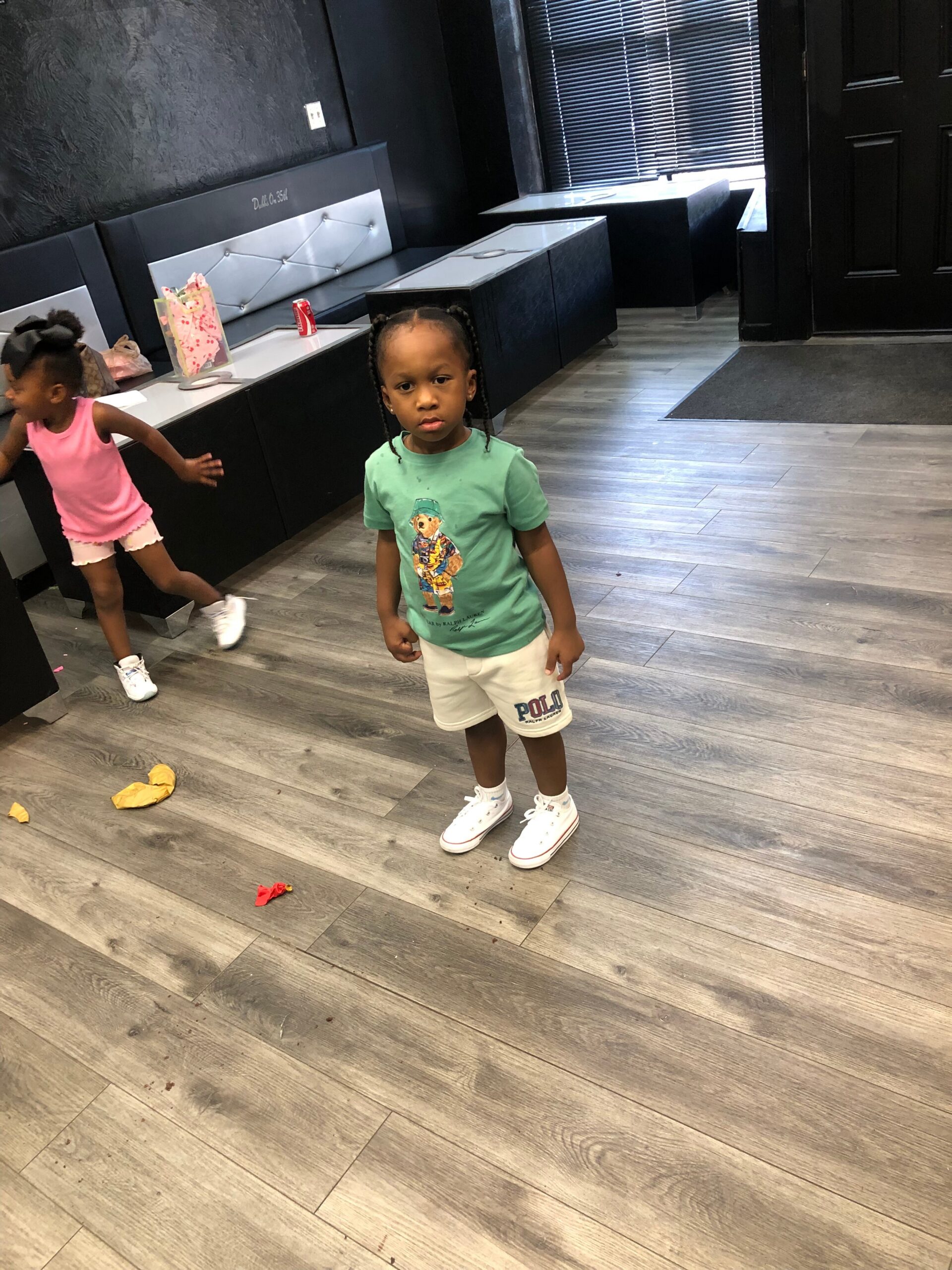 Two young children in a dark, modern room; the girl in a teal shirt and white shorts stands in the foreground while another girl in pink runs along a counter with bags and a red soda can nearby.