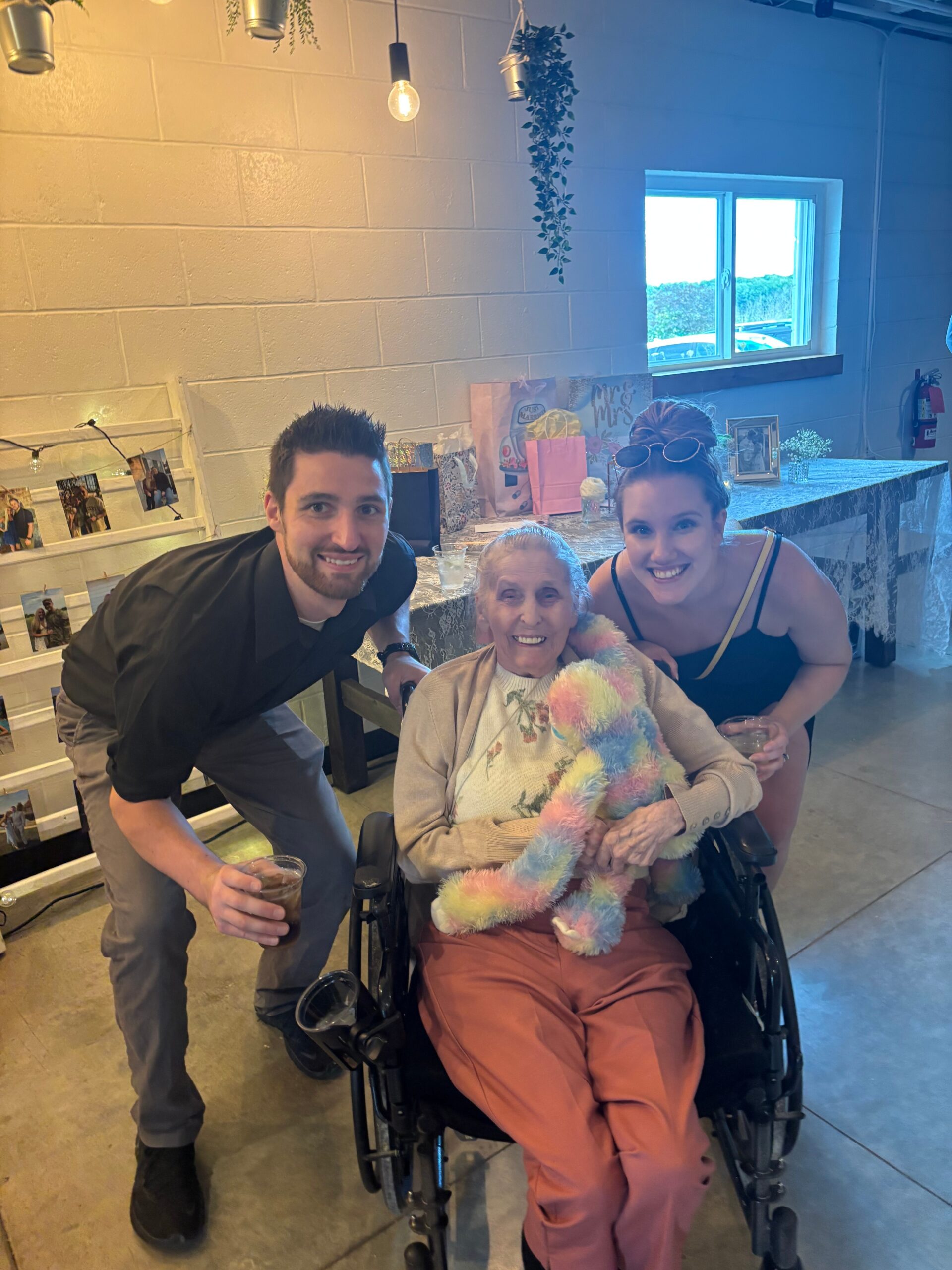 Man kneels beside an elderly woman in a wheelchair, smiling with two younger women at a casual indoor gathering.