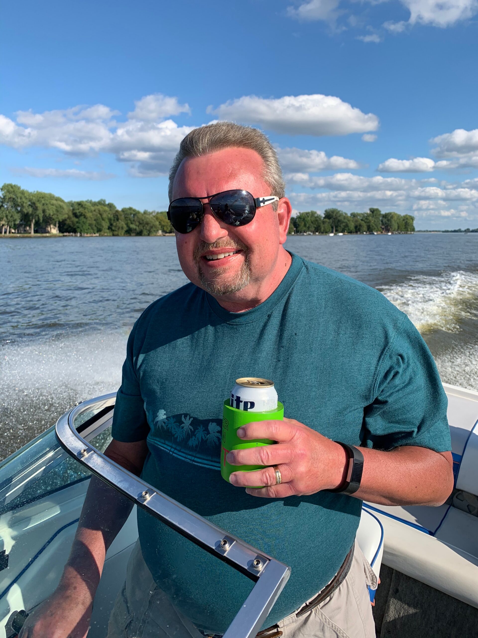 Smiling man in sunglasses on a boat, holding a can in a green koozie with blue sky and water in the background