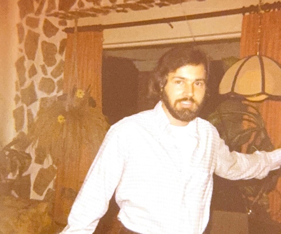 Bearded man in a light checkered shirt leaning against a doorway in a living room with a stone wall, orange curtains, and a domed lamp.